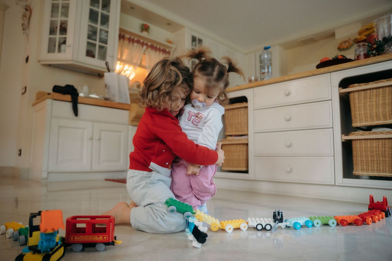 Two young children sharing a sweet hug while playing with colorful toy trains on floor.