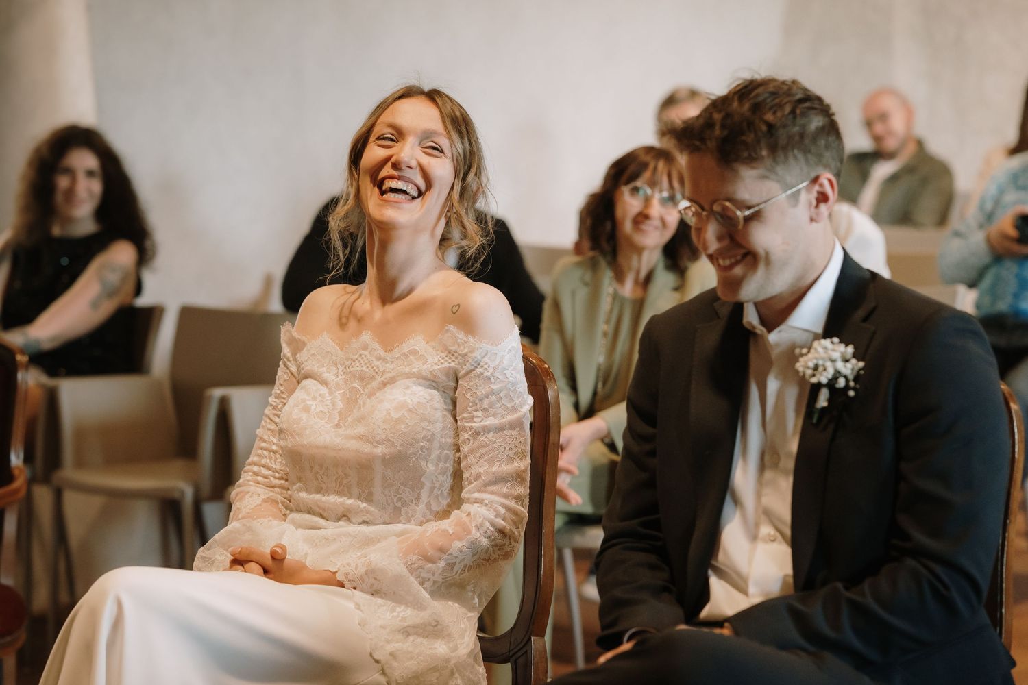 Bride and groom laughing joyfully during wedding ceremony seated in chairs with guests in background.