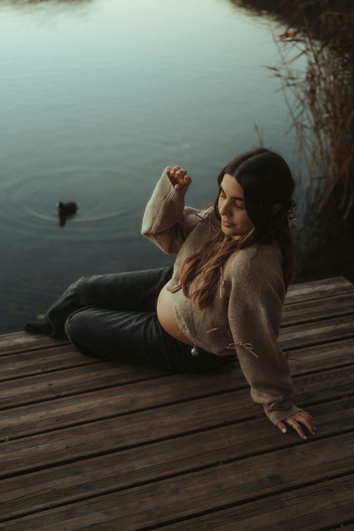 Pregnant woman sitting on wooden dock by calm lake at dusk.