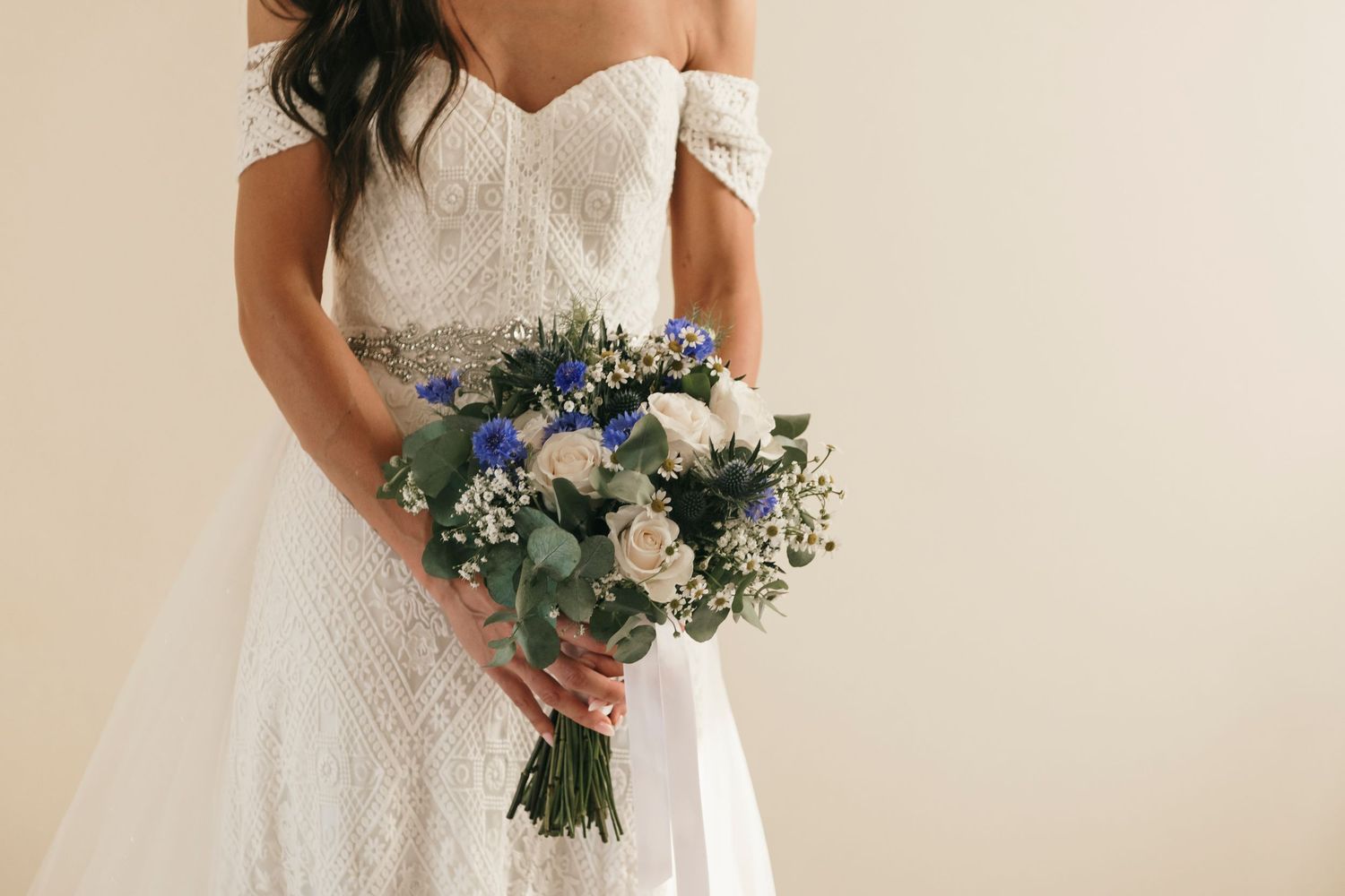 Bride in off-shoulder lace wedding gown holding blue and white floral bouquet with greenery against neutral background.