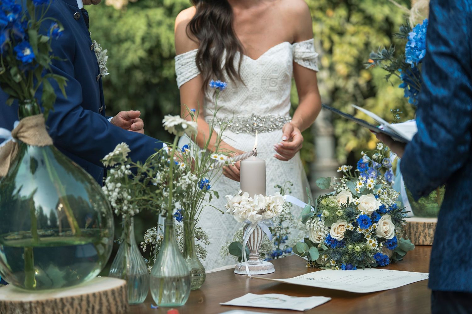 Bride in off-shoulder lace gown stands at altar with blue and white floral arrangements during ceremony.