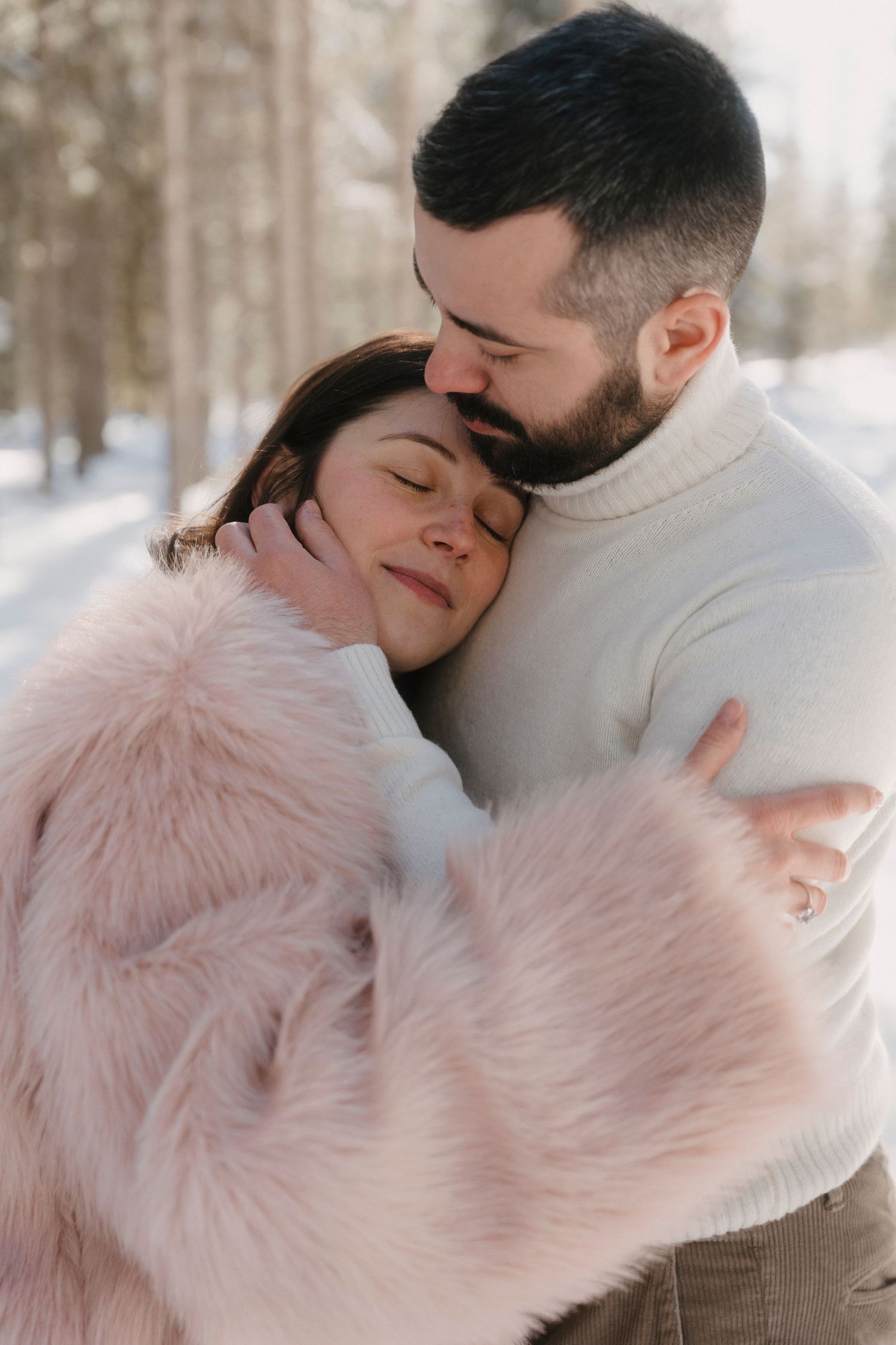 Man in white turtleneck tenderly embracing pregnant woman in pink fur coat in a winter forest setting.