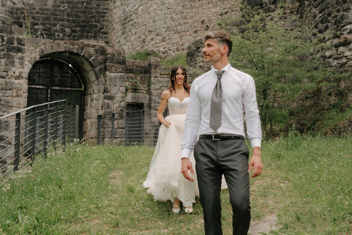 Una sposa in abito bianco e uno sposo in completo camminano vicino ad antiche rovine in pietra durante il loro matrimonio.