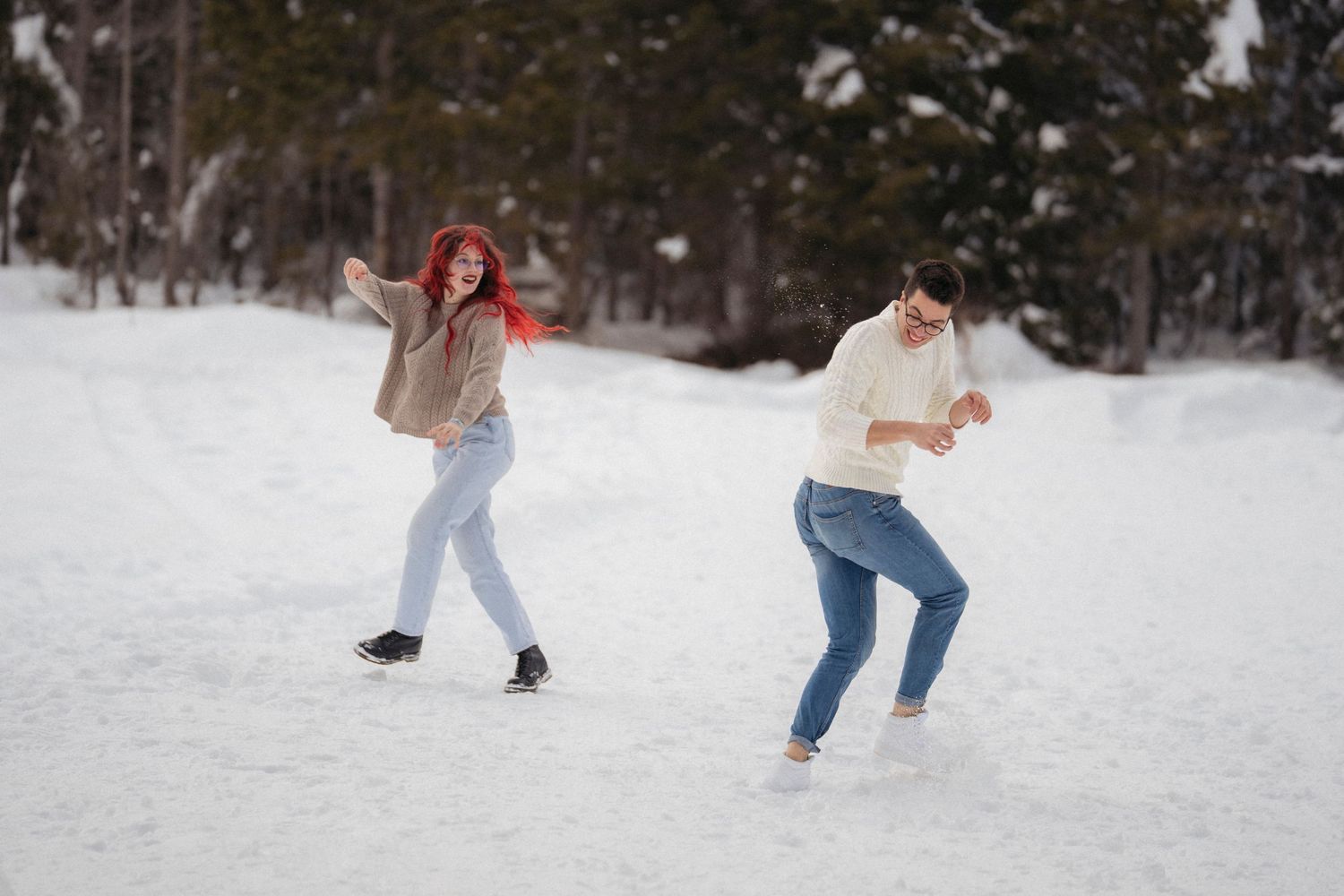 A playful couple dances and throws snowballs at each other on a snowy path surrounded by snow-covered trees in winter.