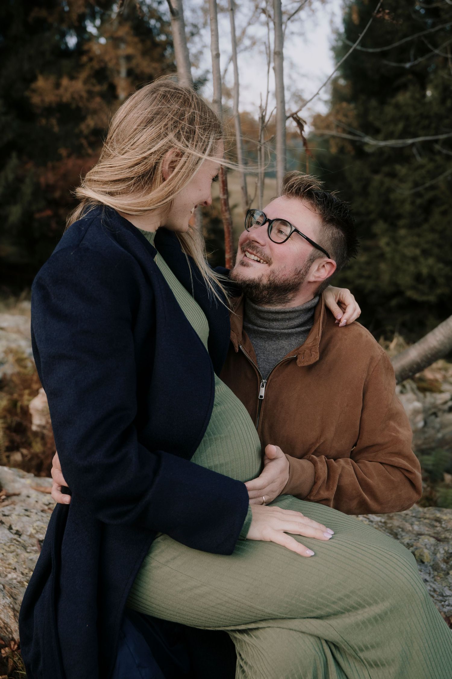 A bearded man lovingly embraces his pregnant partner in an autumn forest setting.
