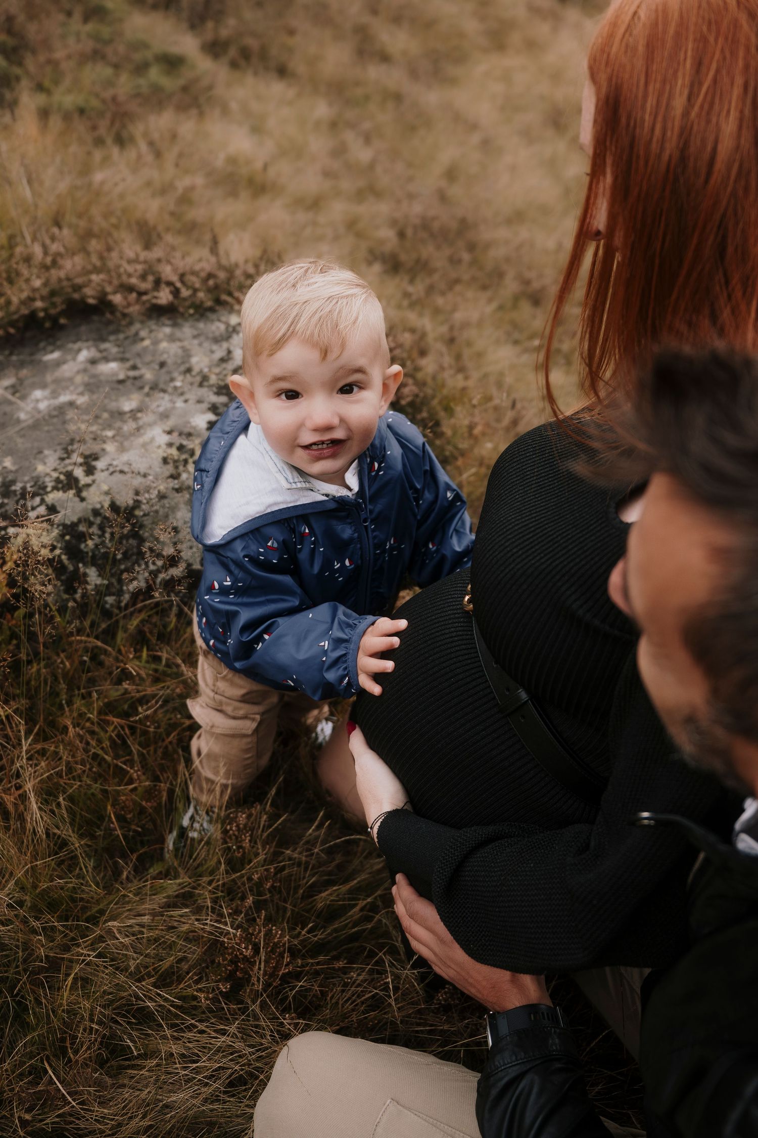 Smiling toddler in blue jacket being supported by parents in an autumn grass field.