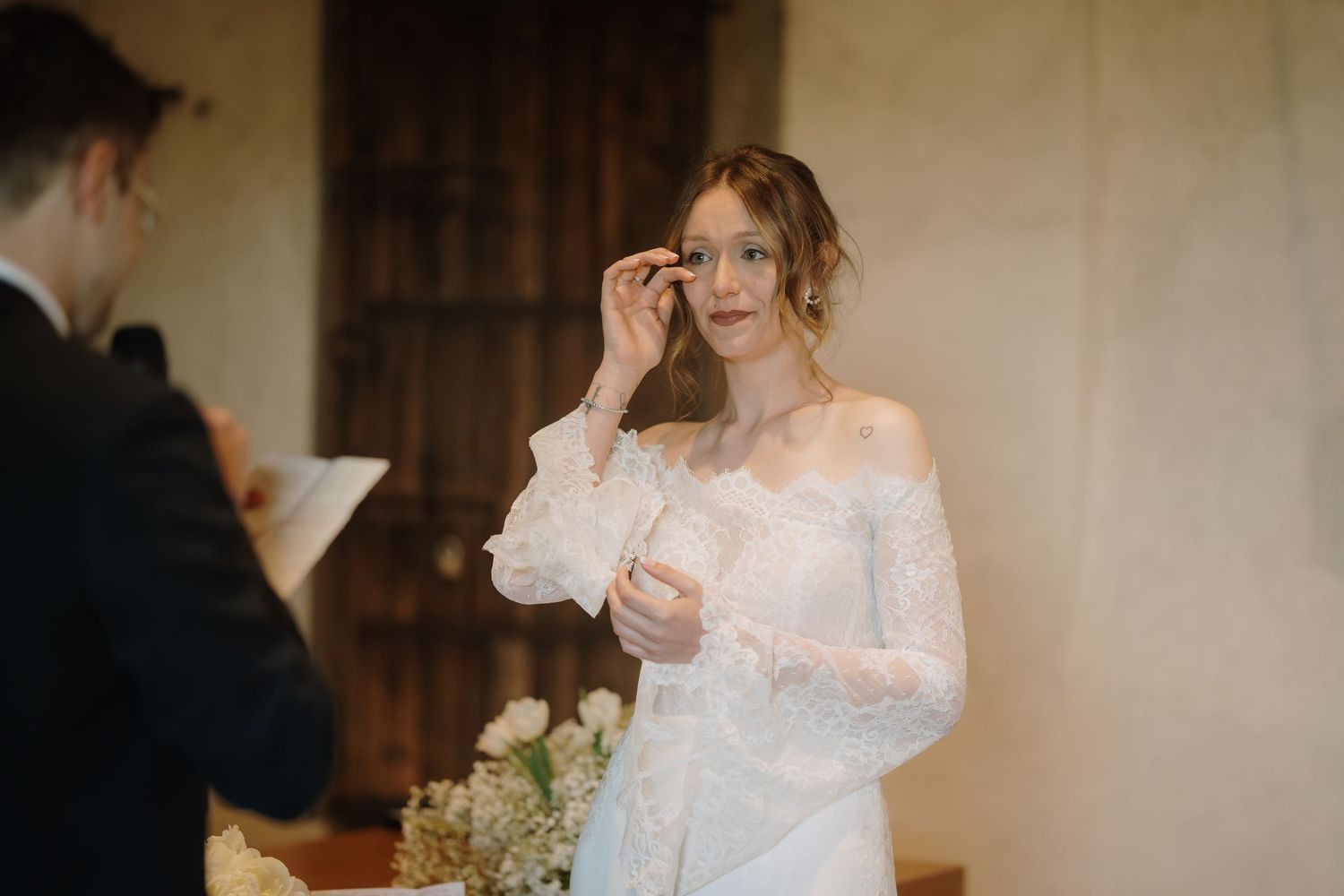 Emotional bride in lace off-shoulder wedding dress wiping tears during heartfelt ceremony vows.