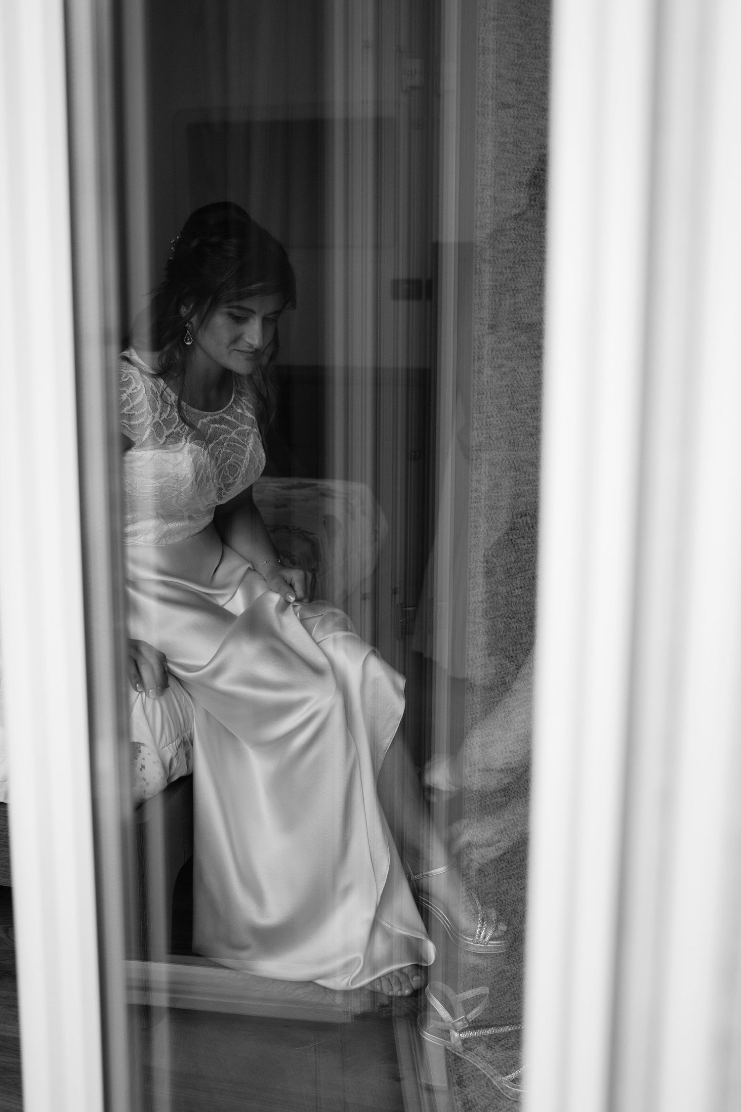 Black and white portrait of bride in elegant lace wedding gown sitting by a window before ceremony.