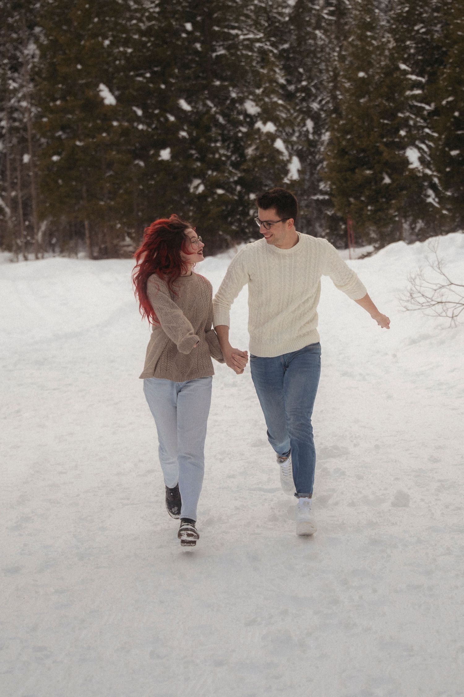 A happy couple holds hands and runs together along a snowy winter path surrounded by snow-covered evergreen trees.
