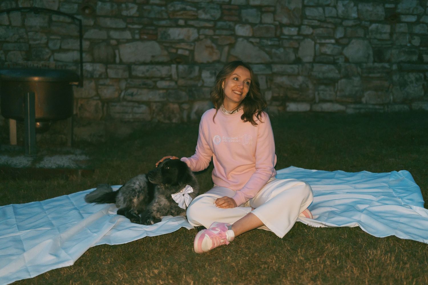 Young woman in pink sweater sitting on blanket outdoors with black dog against stone wall.