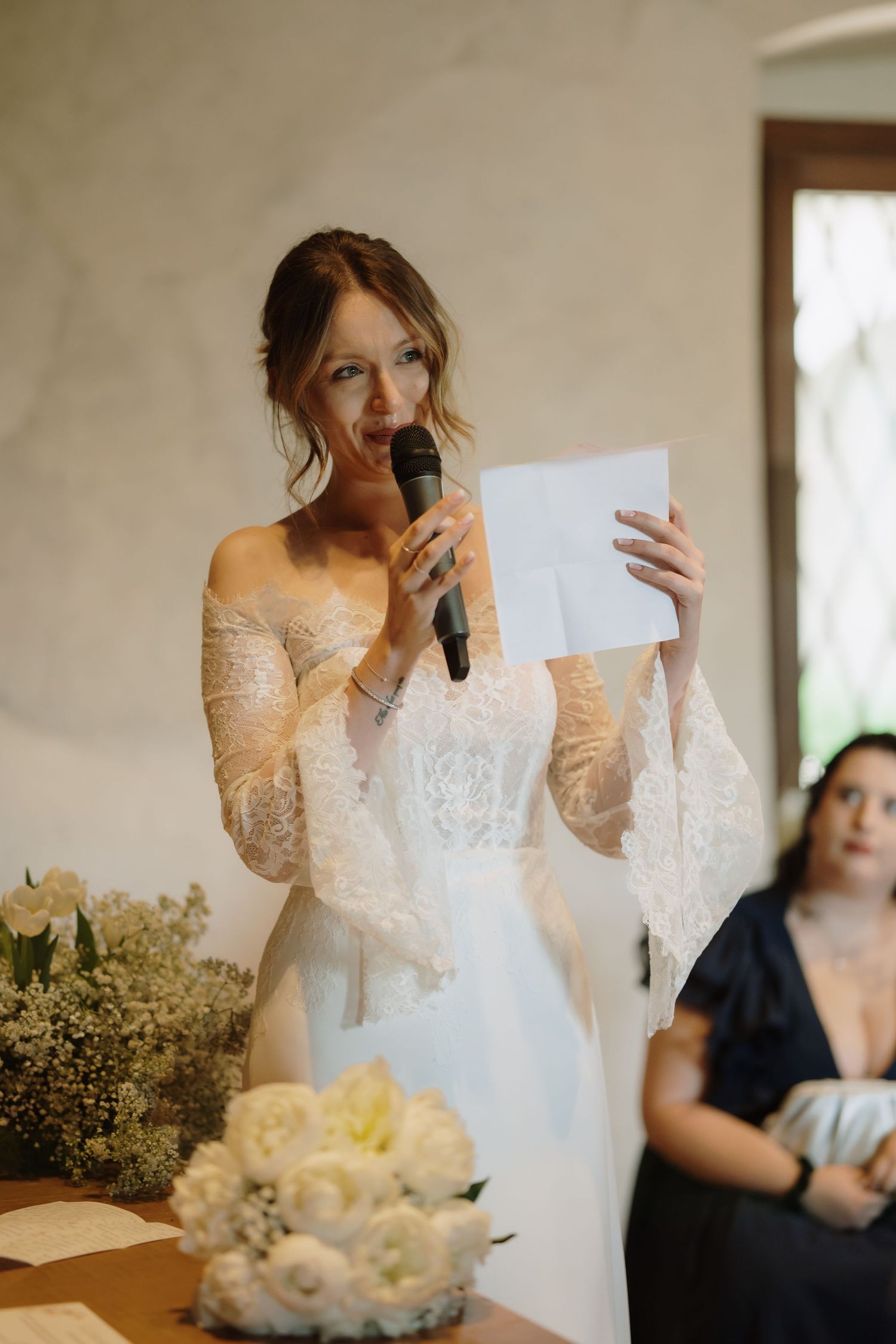 Bride in lace off-shoulder gown reads vows into microphone while holding paper at wedding ceremony.