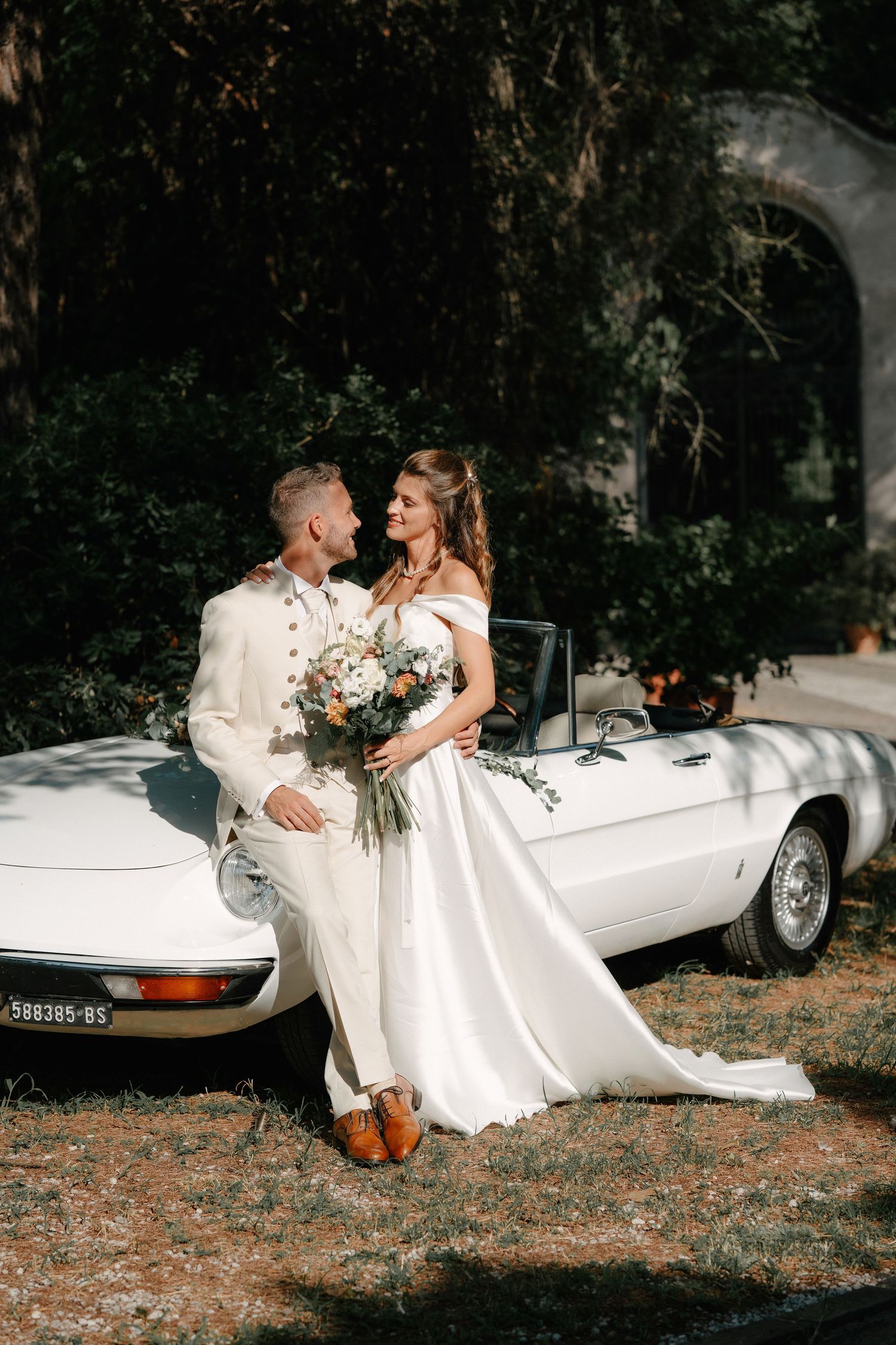 Bride and groom pose together leaning against a white vintage convertible car outdoors on their wedding day.