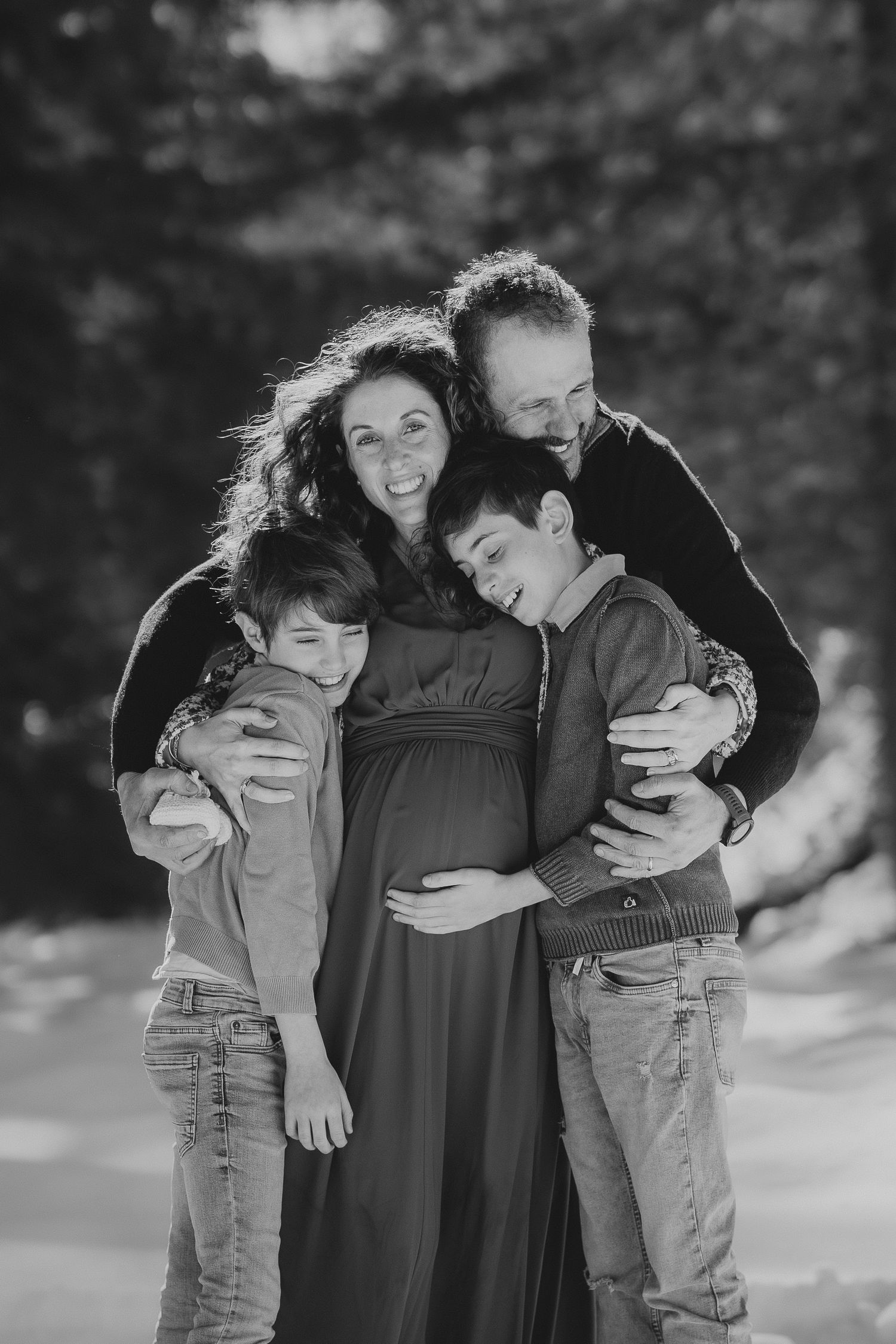 Black and white family portrait with pregnant mother, father, and two sons hugging outdoors.