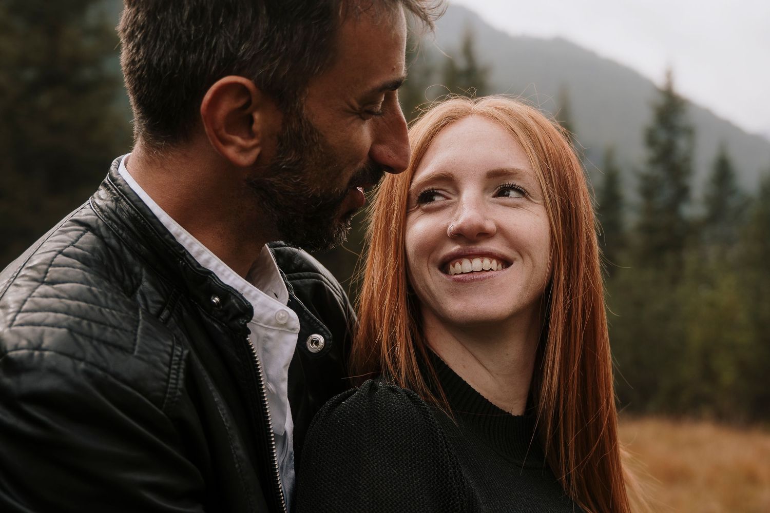 Smiling redhead woman and bearded man sharing a tender moment in a mountain landscape.
