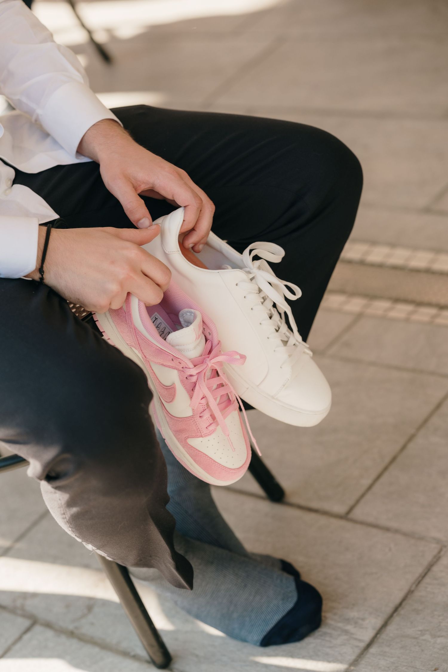 Sposo in camicia bianca tiene in mano scarpe da ginnastica Nike rosa e bianche, seduto all'aperto nel giorno del matrimonio.