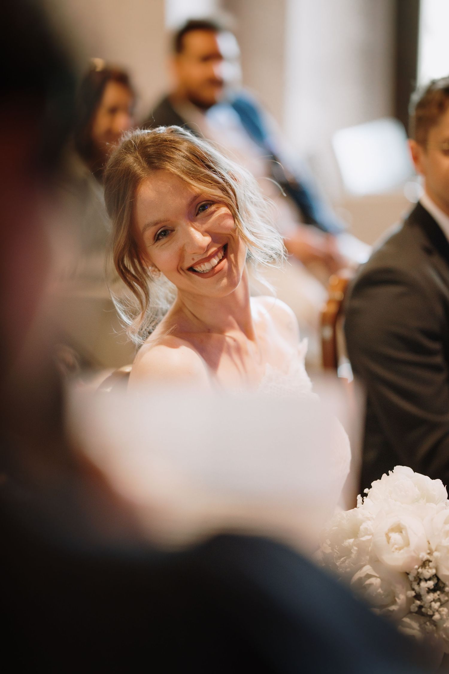 Radiant bride smiling joyfully during wedding ceremony, holding white floral bouquet, with guests visible in background.