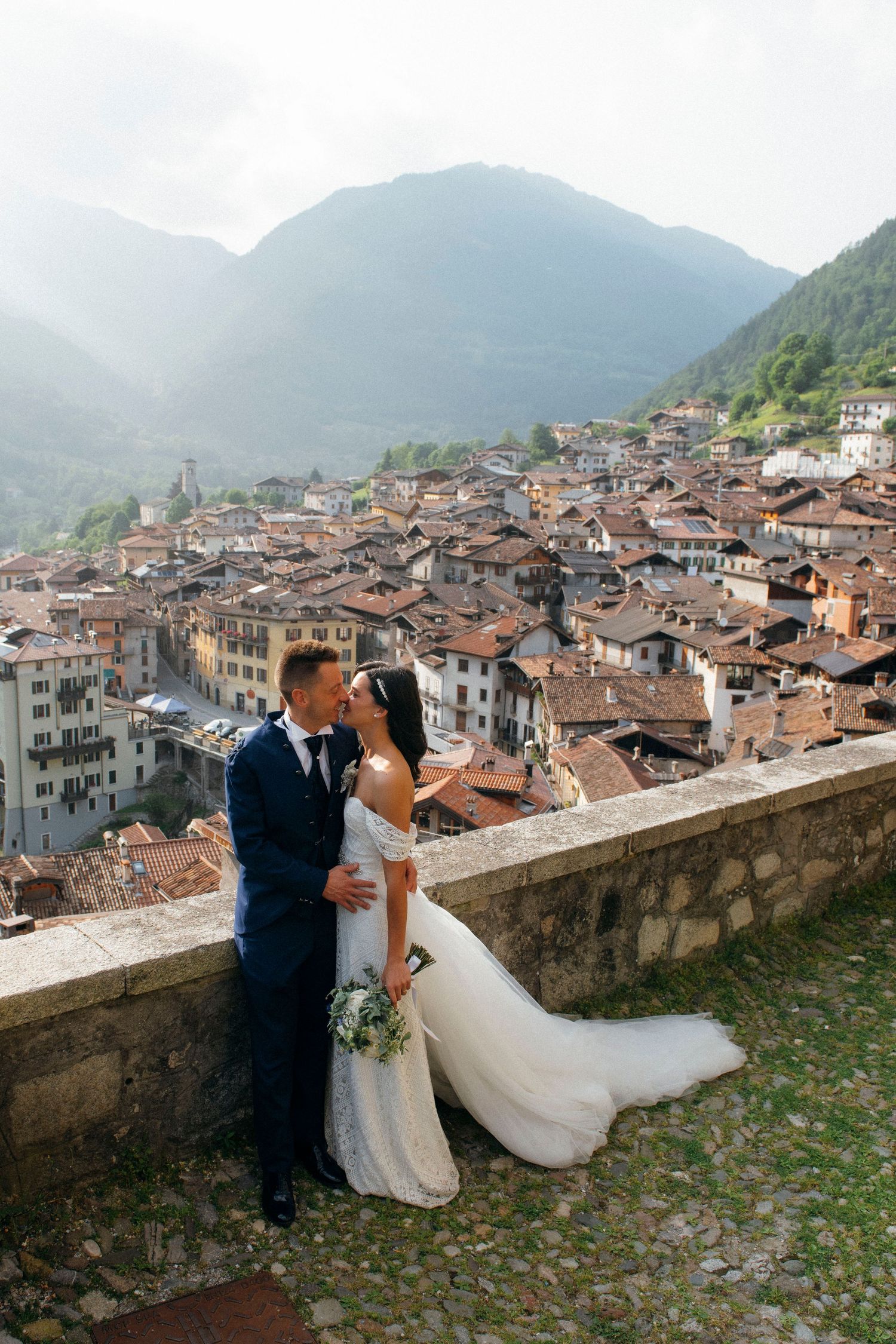 Wedding couple posing on a stone terrace overlooking a picturesque Italian mountain village on their wedding day.