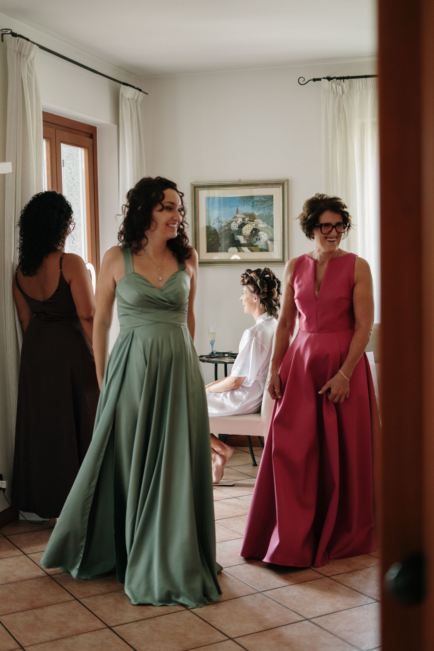 Bridesmaids in green and pink gowns chat indoors while a flower girl sits in the background during wedding preparations.