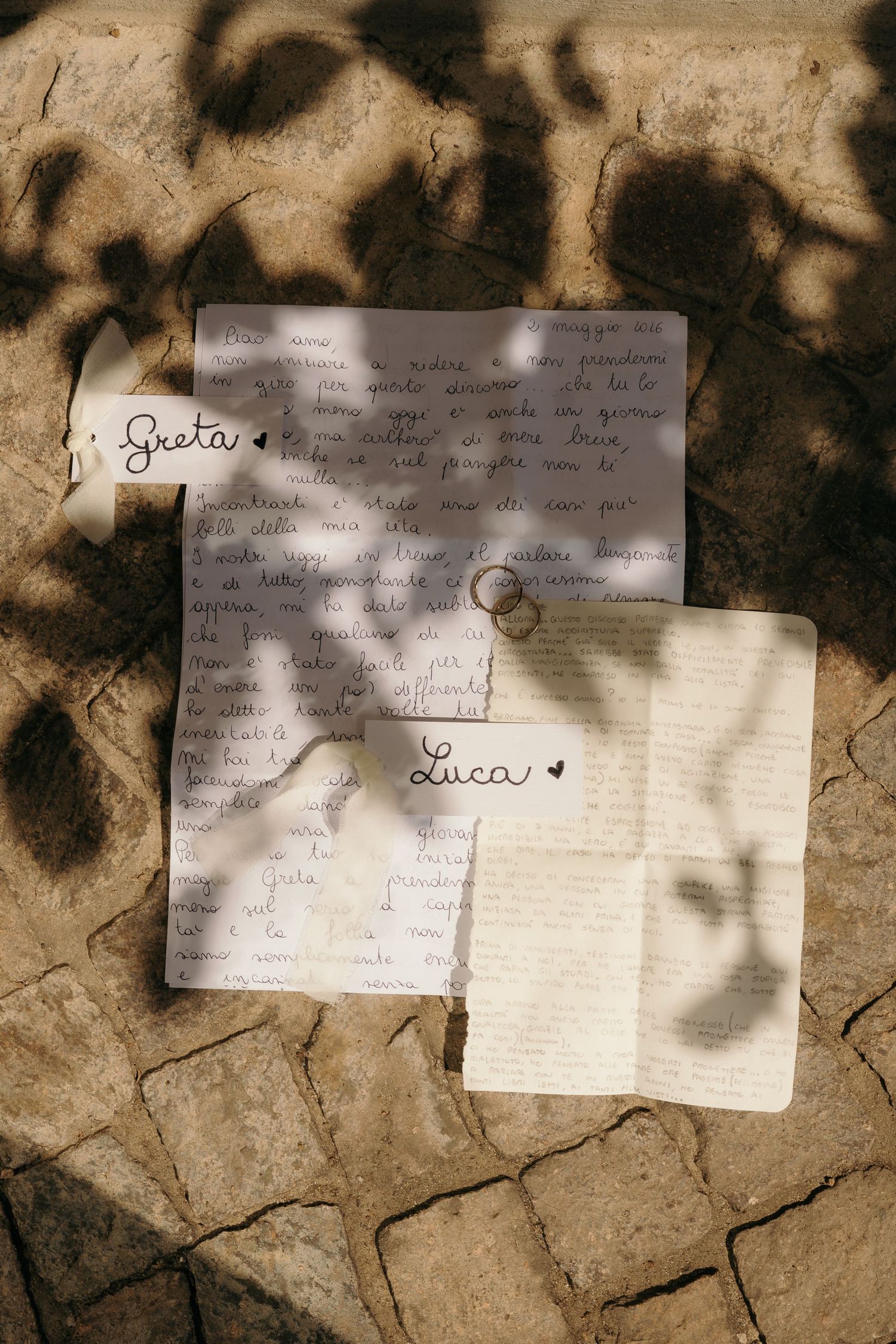 Lettere con le promesse nuziali per Greta e Luca, con fedi d'oro appoggiate su pagine scritte a mano, illuminate da un gioco di luci e ombre