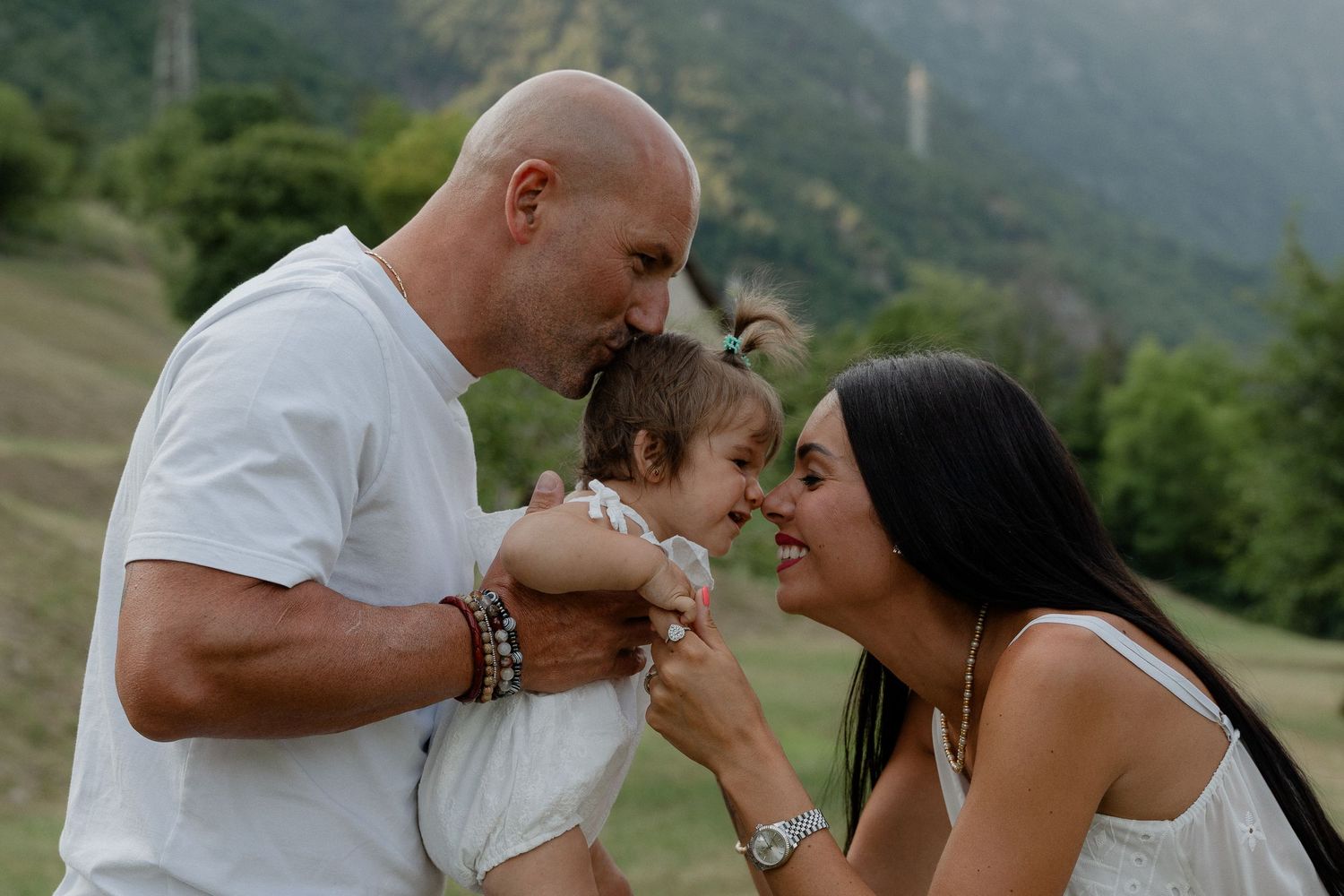 A bald father and dark-haired mother share a tender moment with their baby outdoors in a green mountain setting.