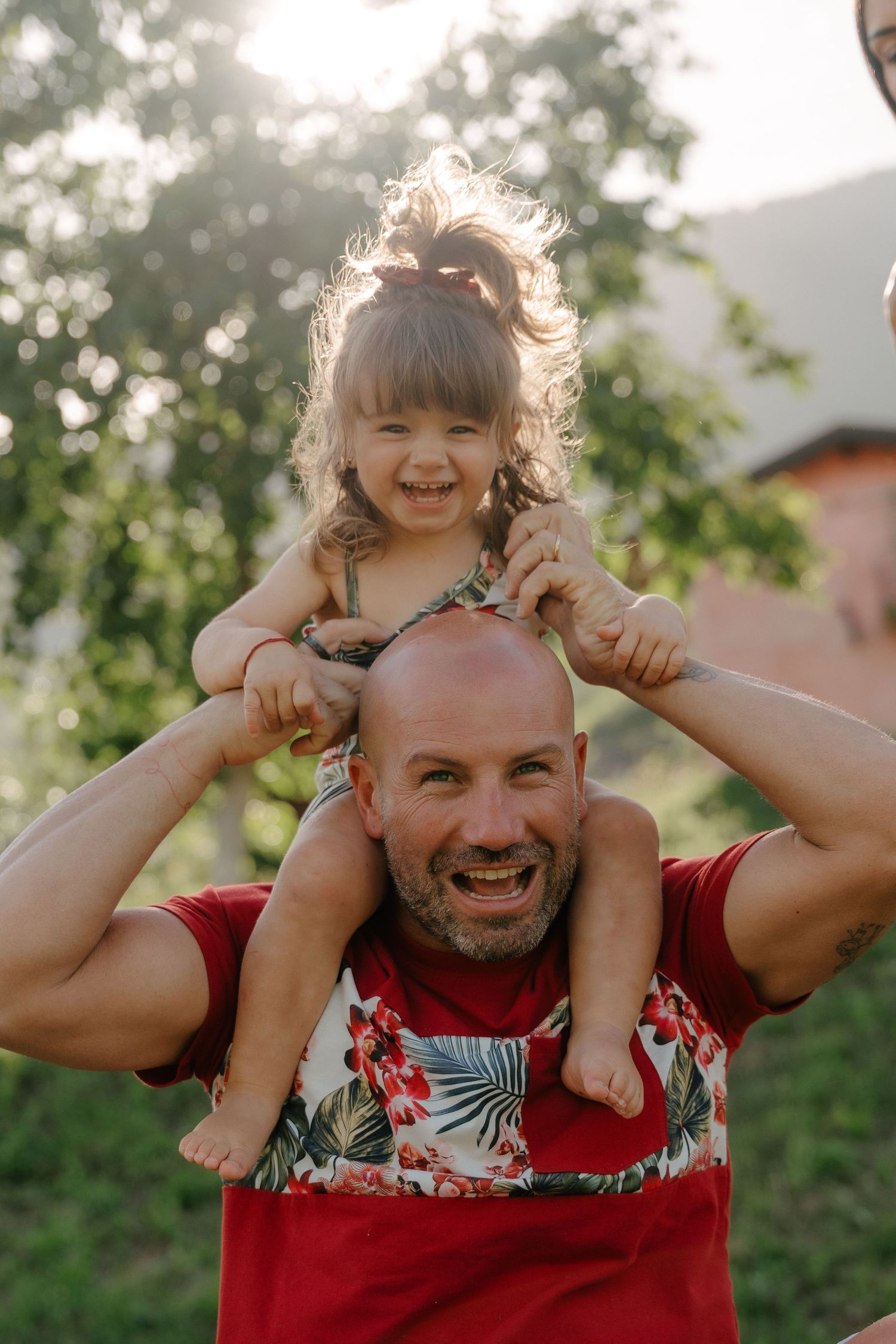 Papà che ride con la figlioletta seduta sulle sue spalle, all'aperto in un parco verde e soleggiato.