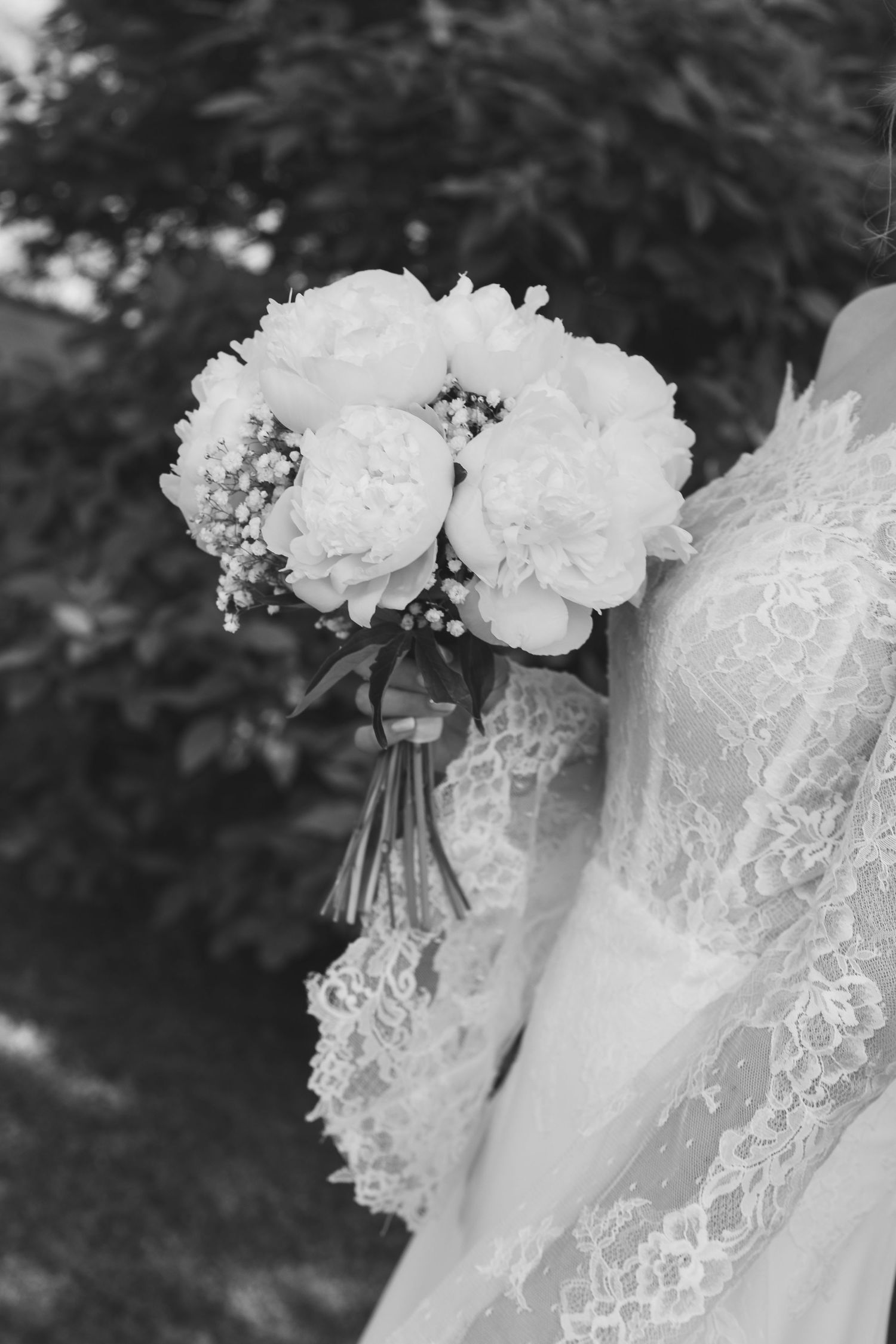 Primo piano in bianco e nero di una sposa che regge un elegante bouquet nuziale di peonie bianche e nebbiolina.