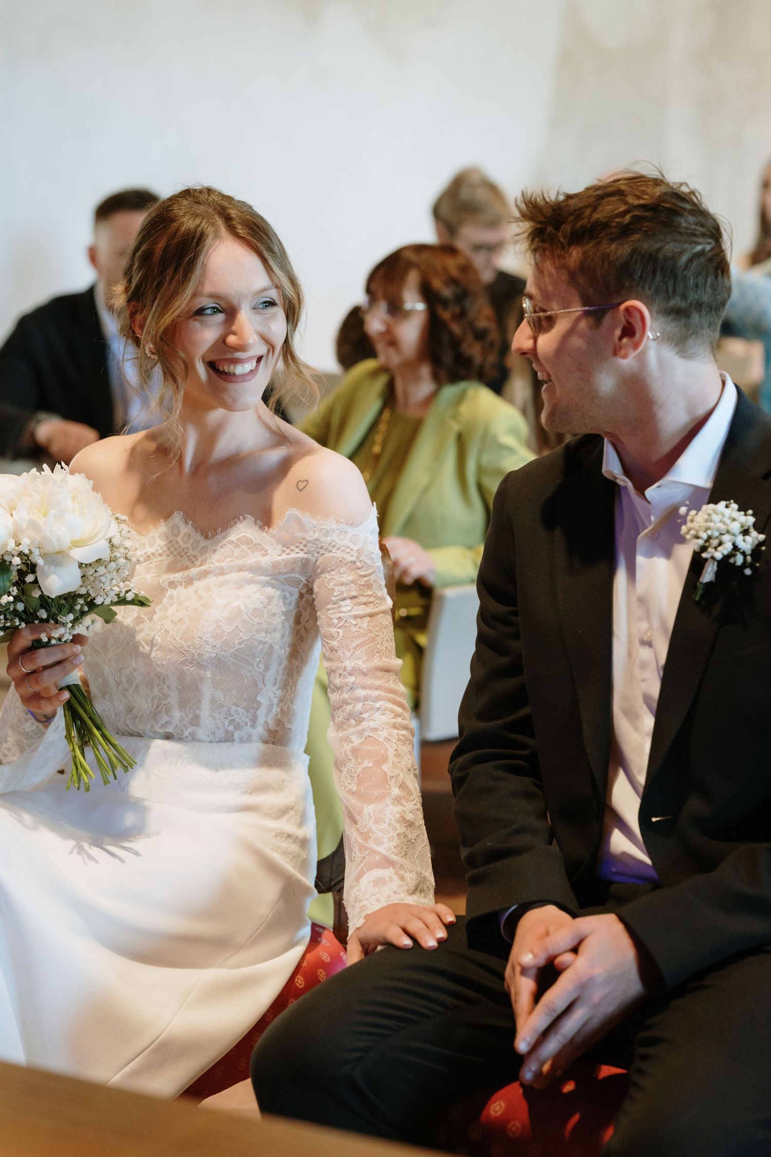 Bride in lace wedding dress laughs joyfully with groom in black suit during wedding ceremony.