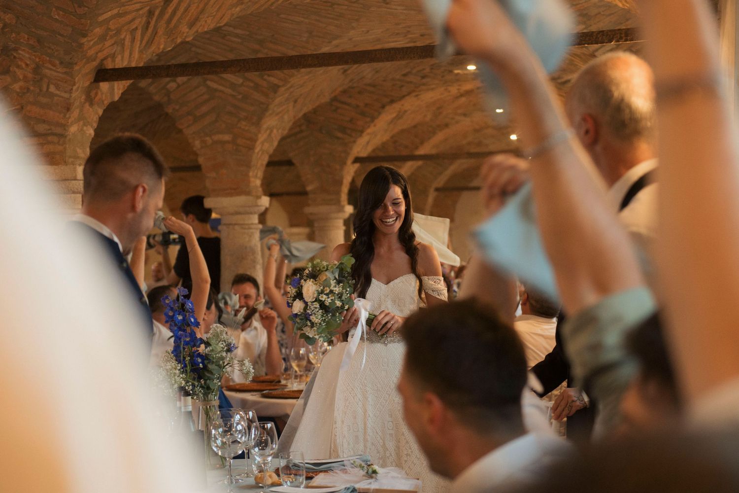 Bride smiling at wedding reception in rustic vaulted brick venue with guests raising hands in celebration.