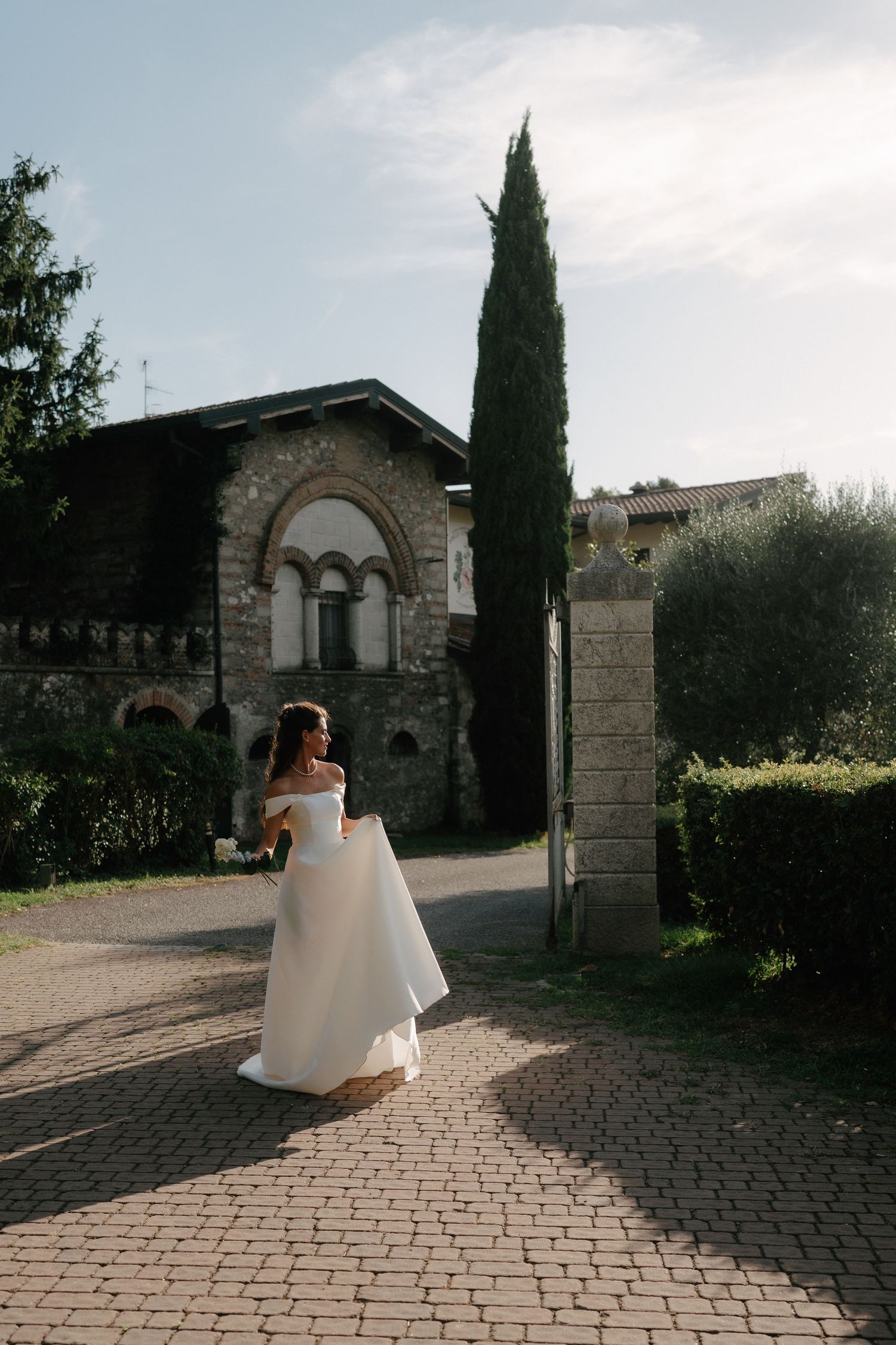 Bride in white off-shoulder gown walks cobblestone path by stone villa with cypress tree in sunlit garden.