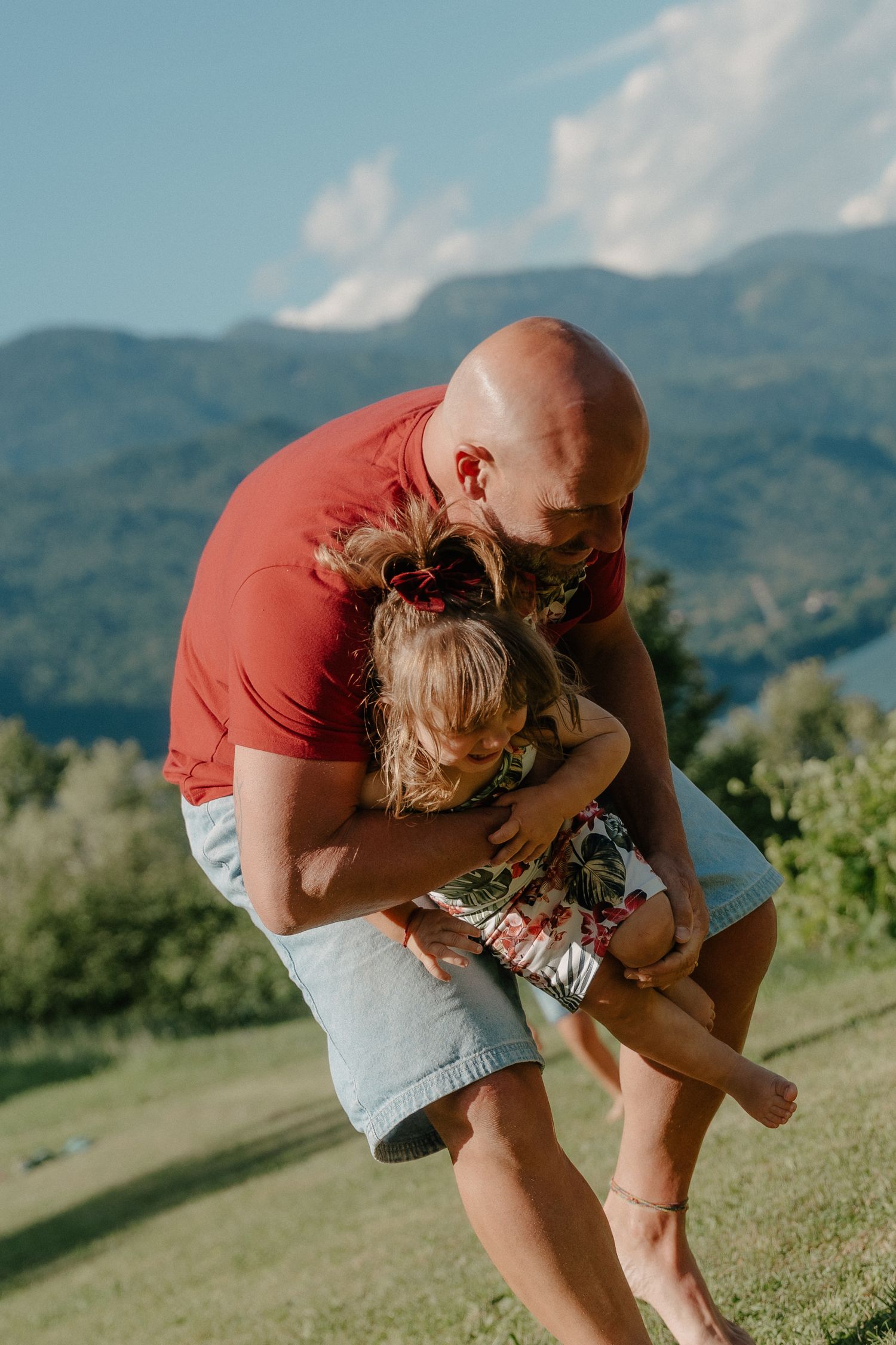 Padre abbraccia scherzosamente la giovane figlia all'aperto, con montagne verdi e cielo azzurro sullo sfondo.