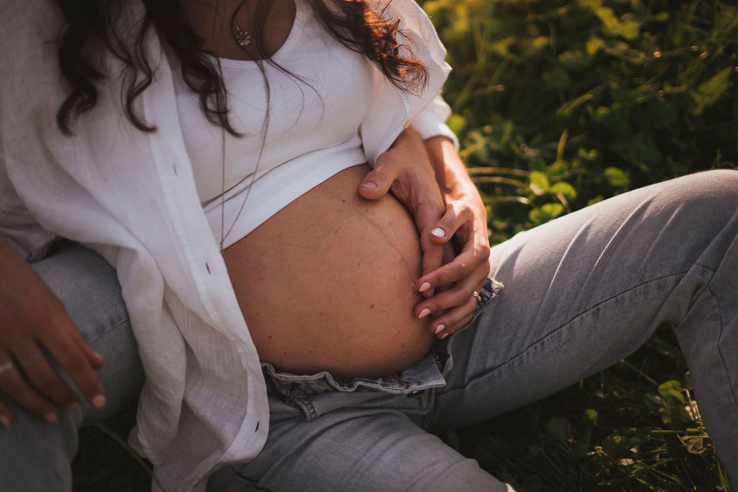 Close-up of a pregnant woman cradling her bare baby bump while sitting outdoors in nature.