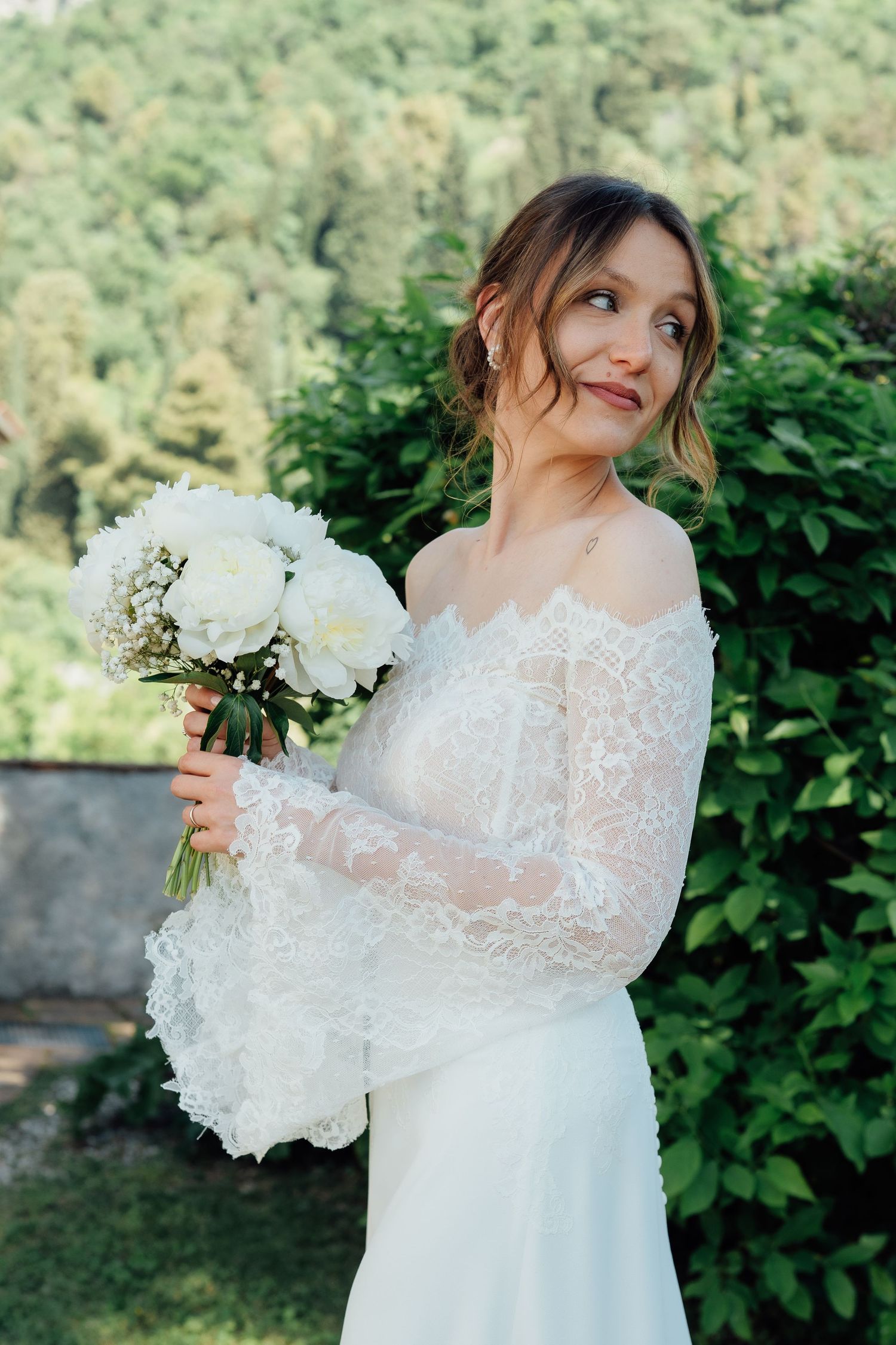 Sposa in abito di pizzo con spalle scoperte regge un bouquet di peonie bianche all'aperto, con uno sfondo di vegetazione rigogliosa.