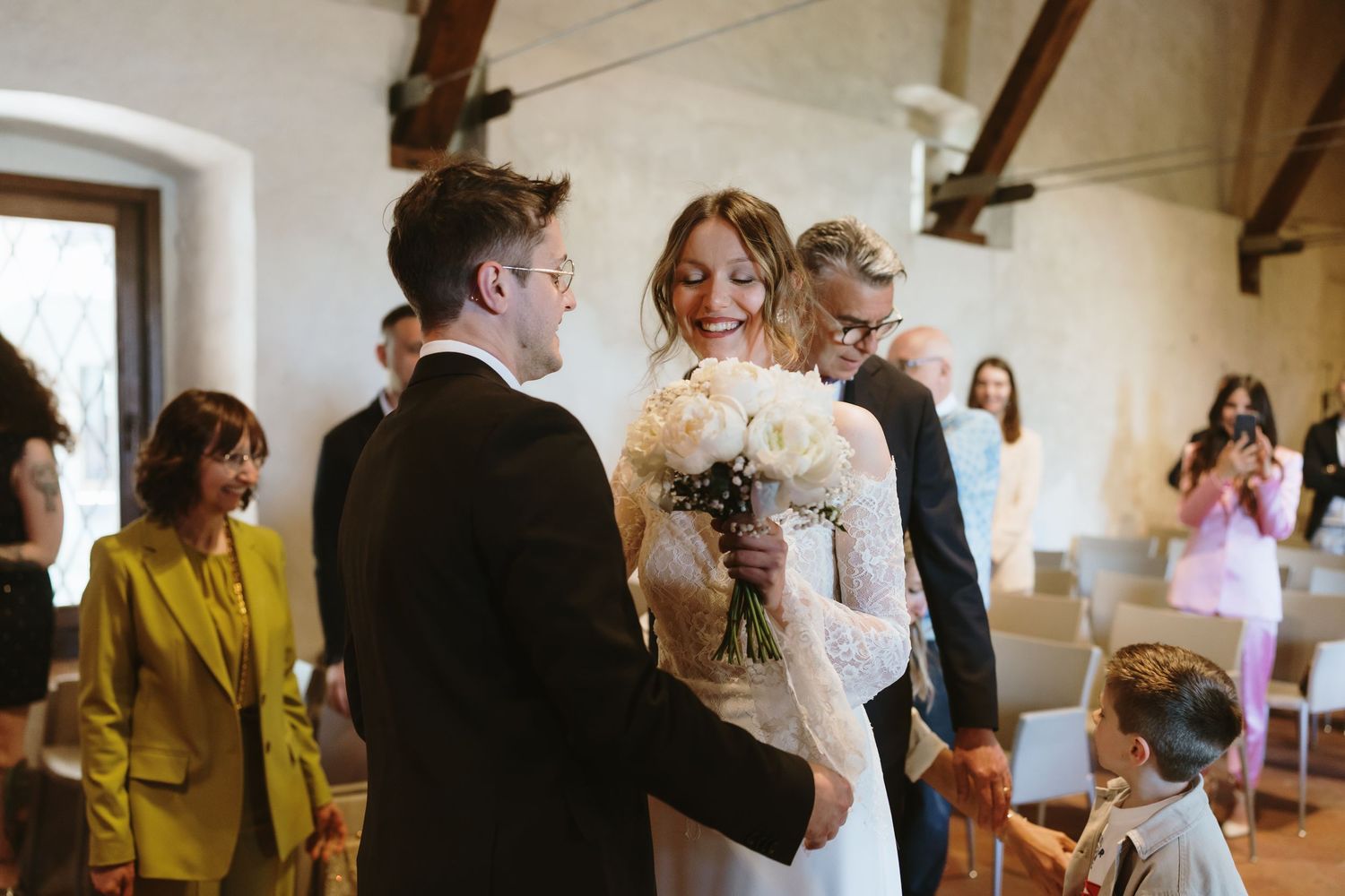 Smiling bride holding white bouquet greets groom at wedding ceremony in elegant stone venue.