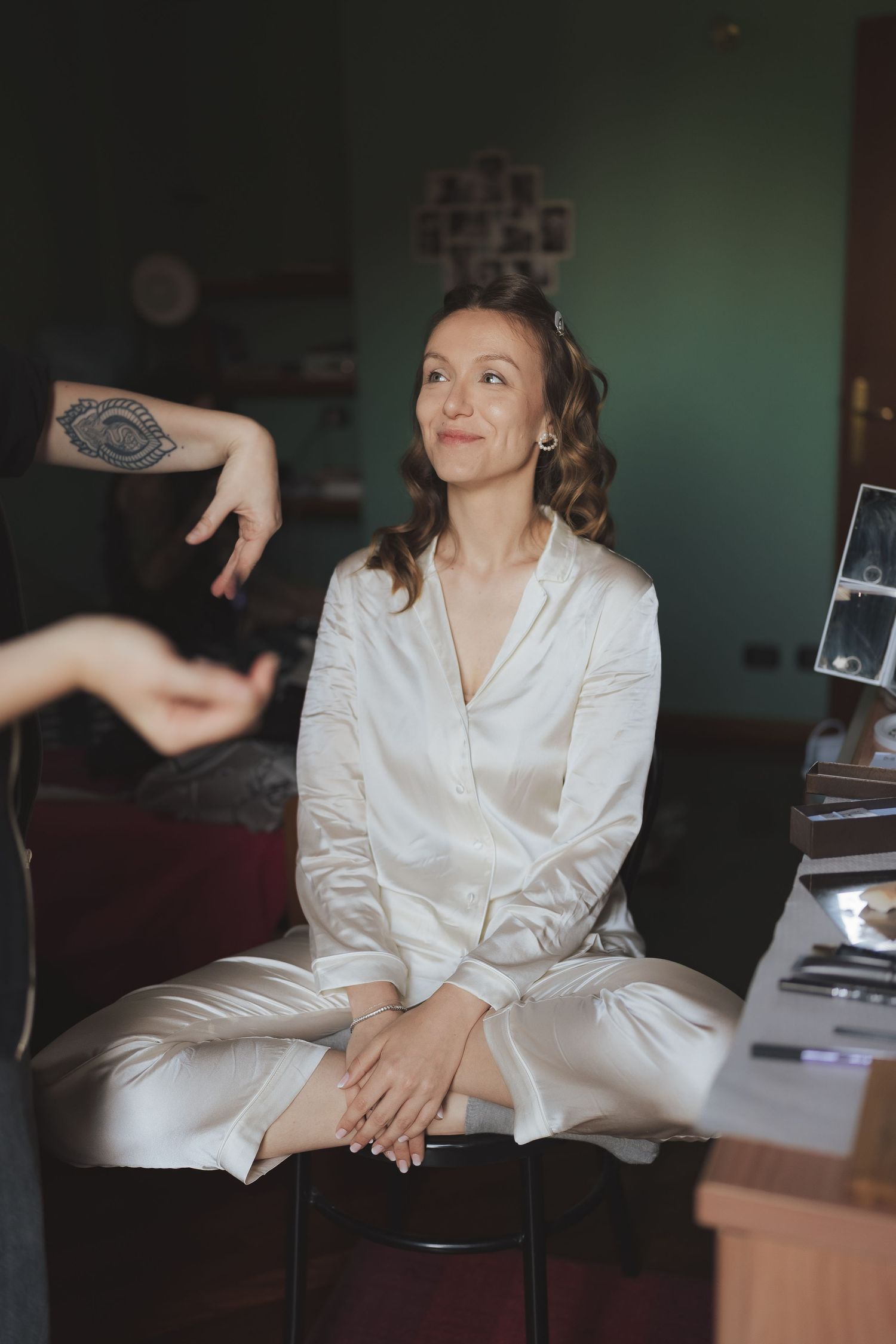 Bride in white satin robe sits on stool getting ready for wedding day with makeup artist nearby.