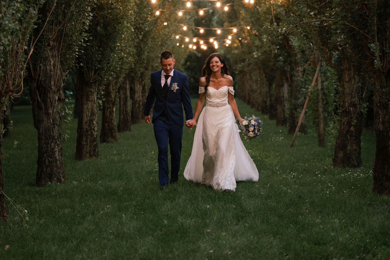 Bride and groom stroll through romantic tree-lined path with string lights at dusk.