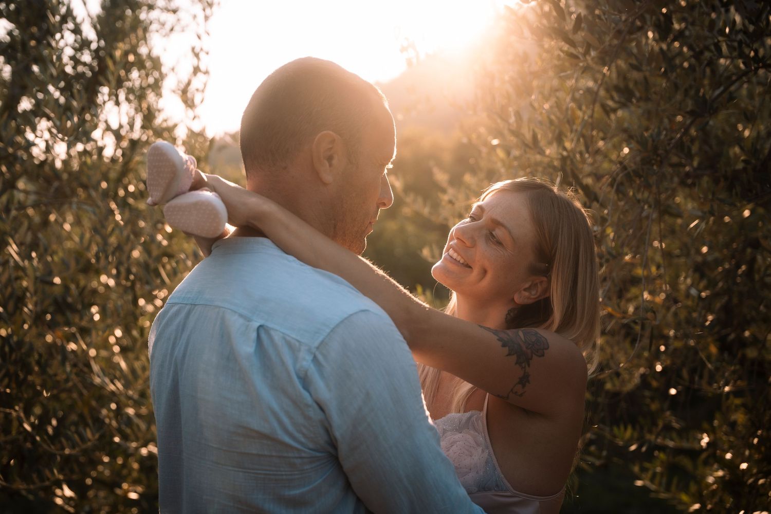 A smiling couple embraces romantically in a sunlit olive grove during golden hour.