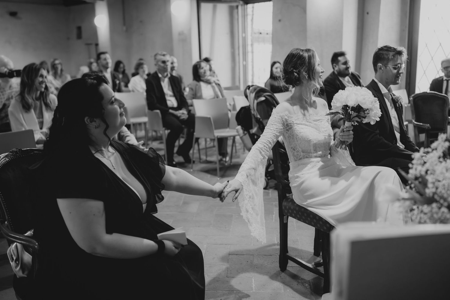 Black and white photo of bride holding hands with a guest while seated during wedding ceremony.