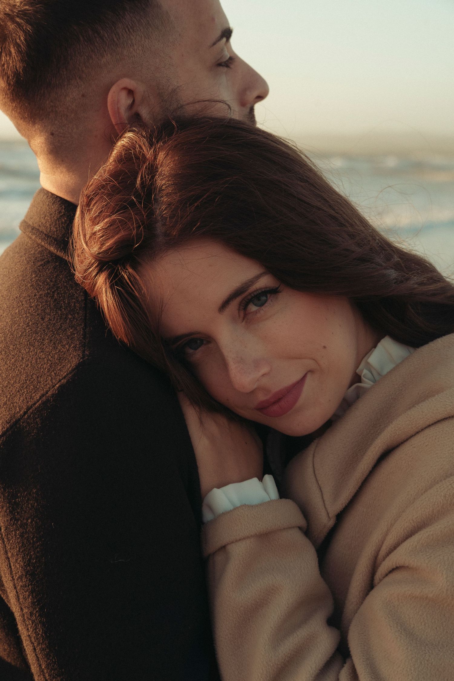 Beautiful woman resting her head on her partners shoulder near the ocean at sunset.