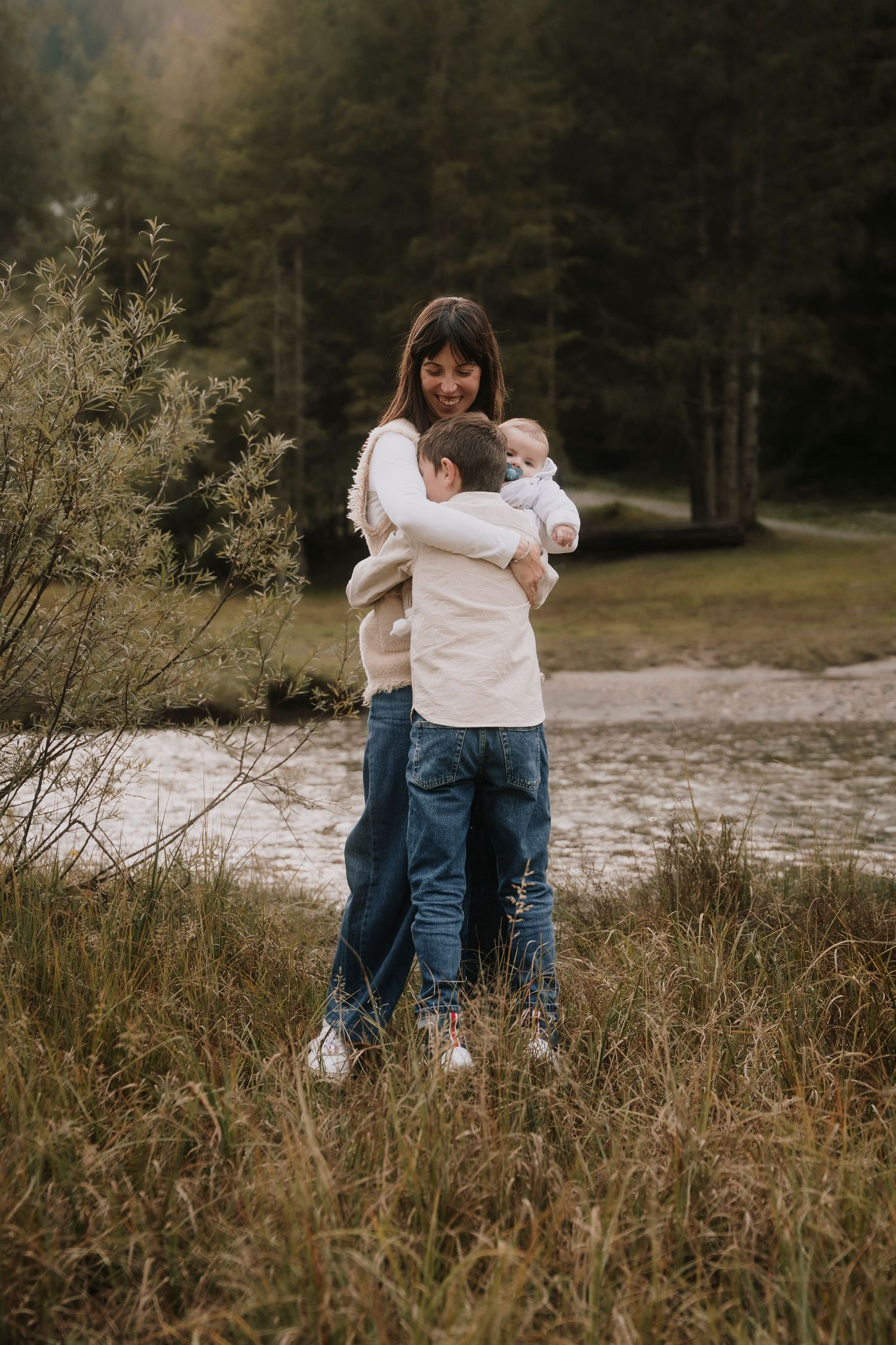 A smiling mother holds her baby while her young son hugs her in a lush outdoor setting.