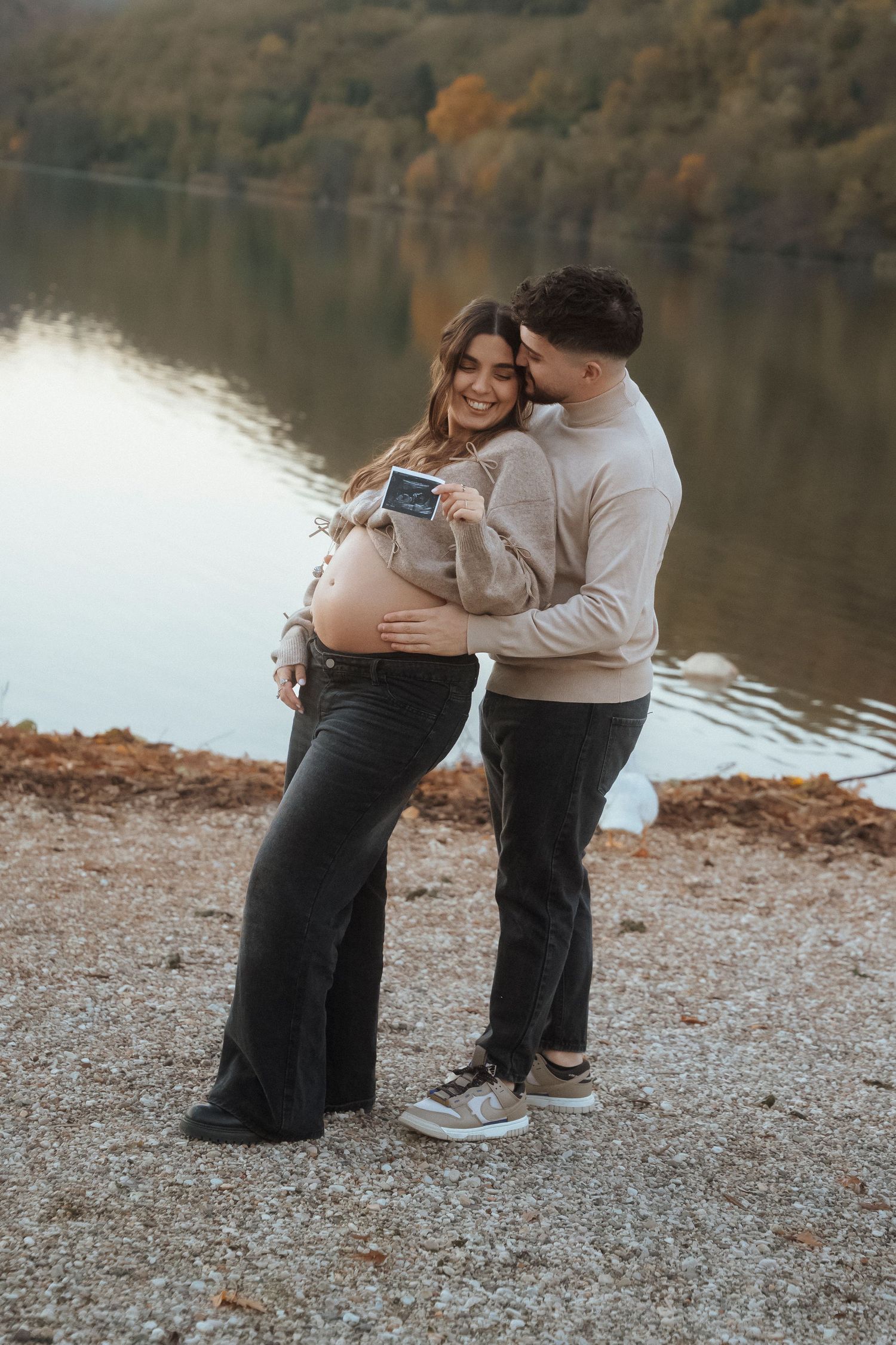 Happy pregnant couple holding ultrasound photo by autumn lake shore together.