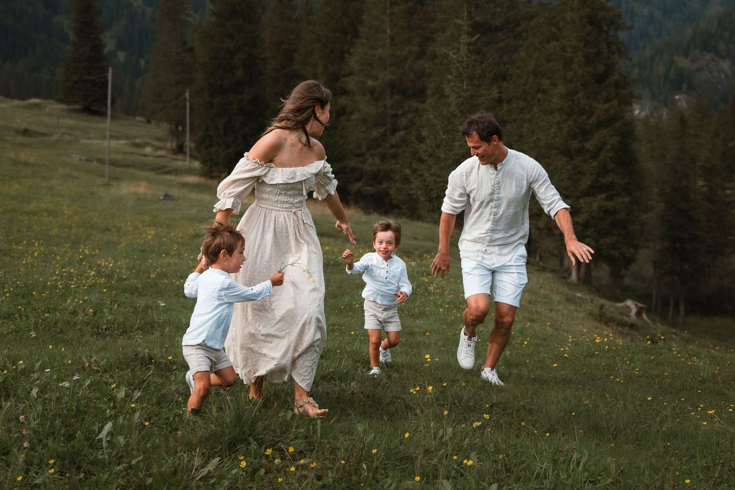 A joyful family with two young children runs together on a green hillside meadow with wildflowers and trees.