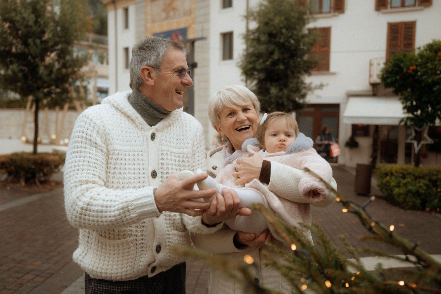 Cheerful grandparents holding a baby near a decorated Christmas tree in an outdoor town square setting.