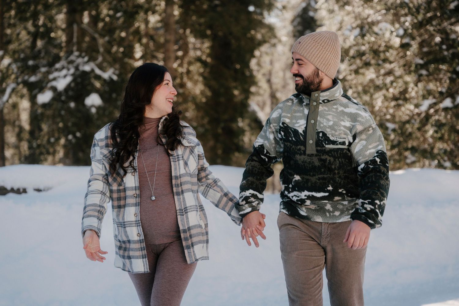 Couple holding hands and smiling at each other in a snowy winter forest, woman showing a baby bump.