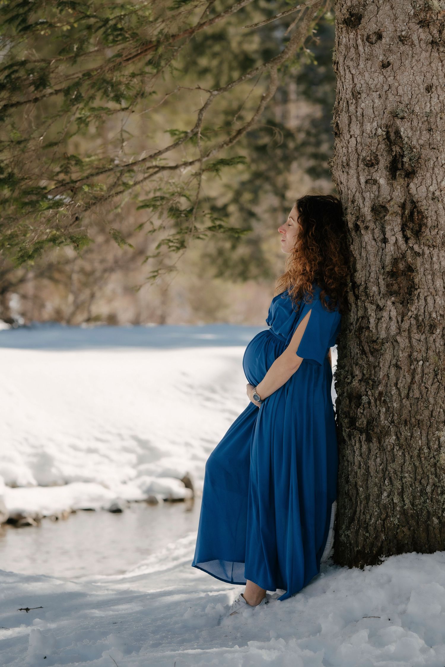 Pregnant woman in blue dress leaning against tree in snowy winter landscape.