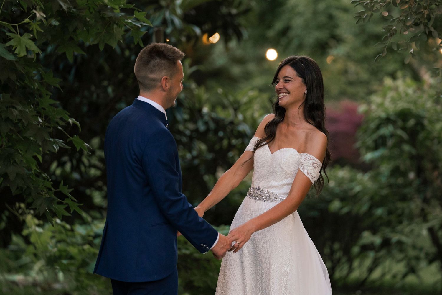 Bride and groom hold hands smiling at each other in lush green garden at twilight.
