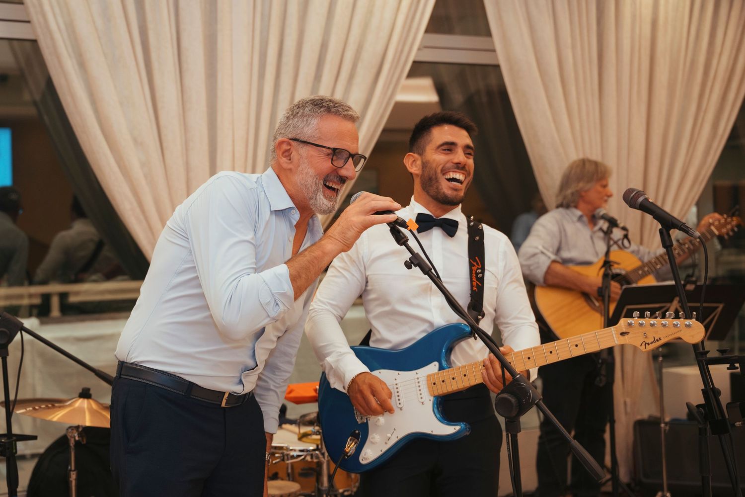 Two men laugh and sing together at a wedding reception while a guitarist performs on stage behind them.