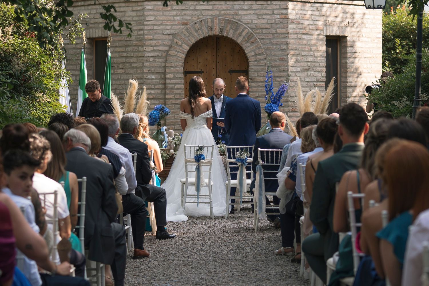 Outdoor wedding ceremony with guests seated in rows as couple stands before officiant at rustic stone building.