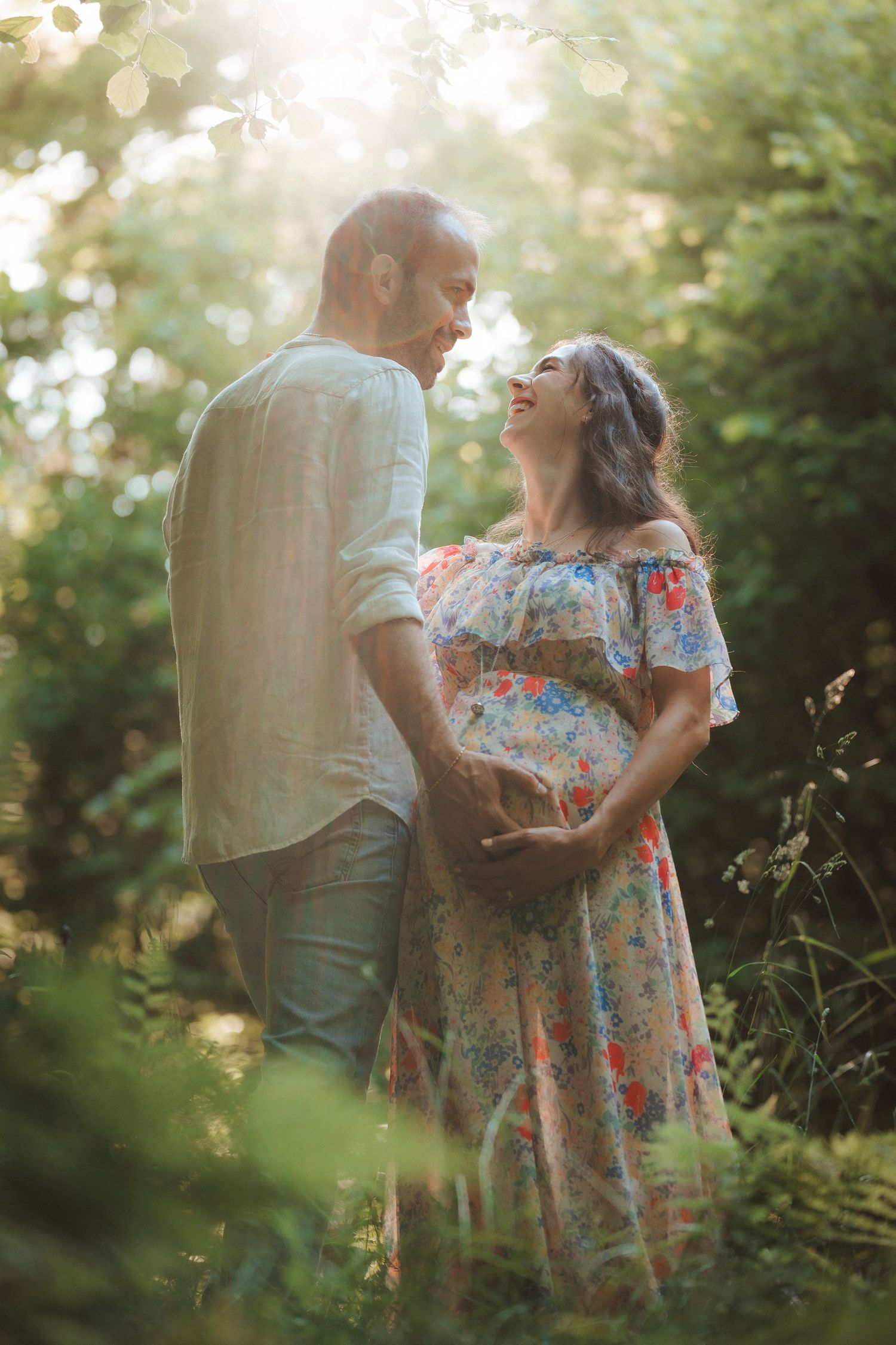 Smiling pregnant couple holding hands and gazing at each other in a sun-drenched garden setting.