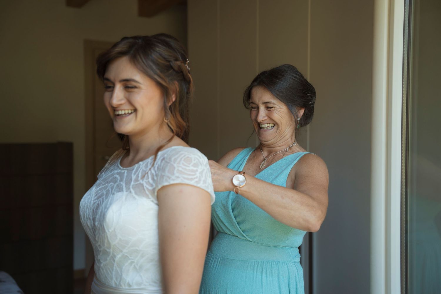 Mother helping bride fasten her white lace wedding dress while both smile joyfully before the ceremony.
