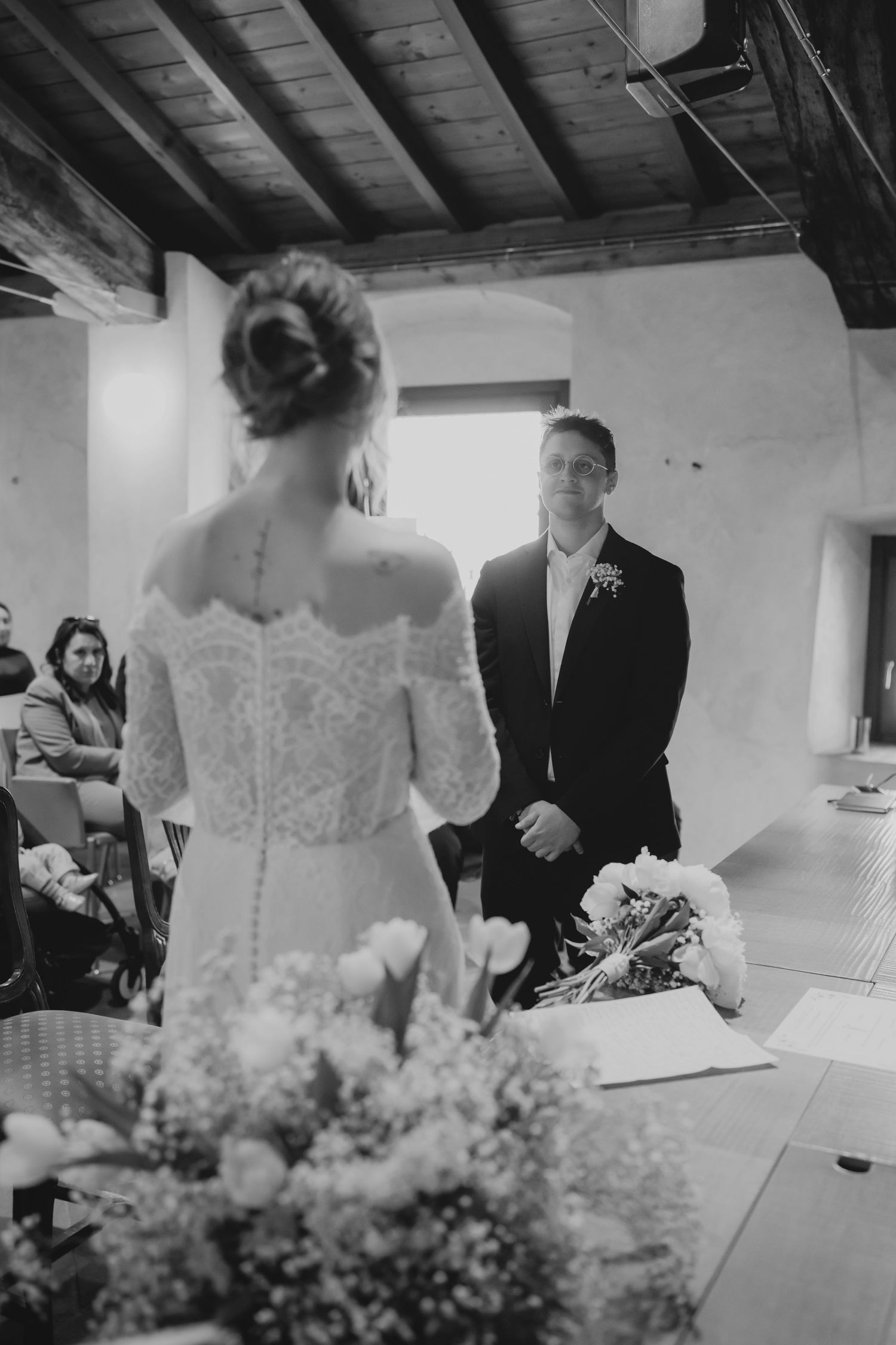 Foto in bianco e nero dello sposo che osserva la sposa avvicinarsi durante una cerimonia nuziale intima e rustica.